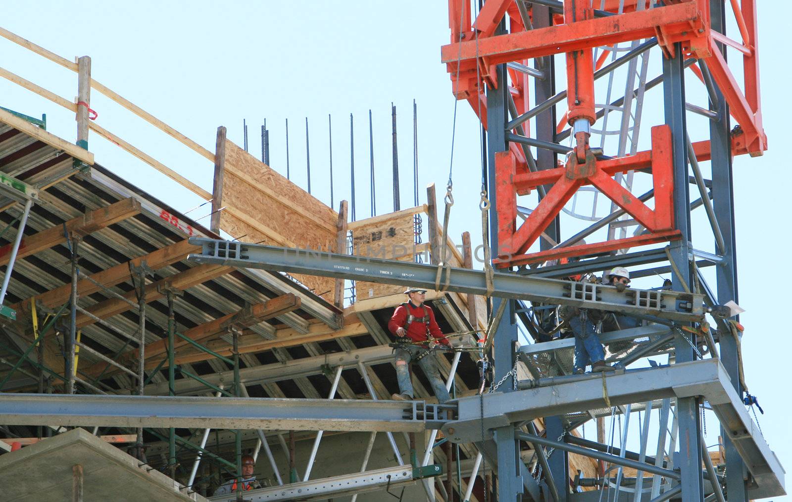 Two construction workers are tethered to the side of a crane that is being erected. They are moving a large steel beam into place.