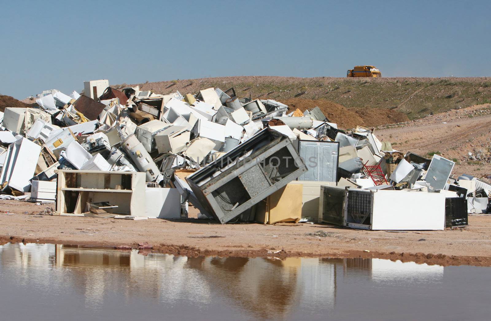 A pile of water heaters, air conditions, and other appliances at a waste dump. A large puddle reflects the image