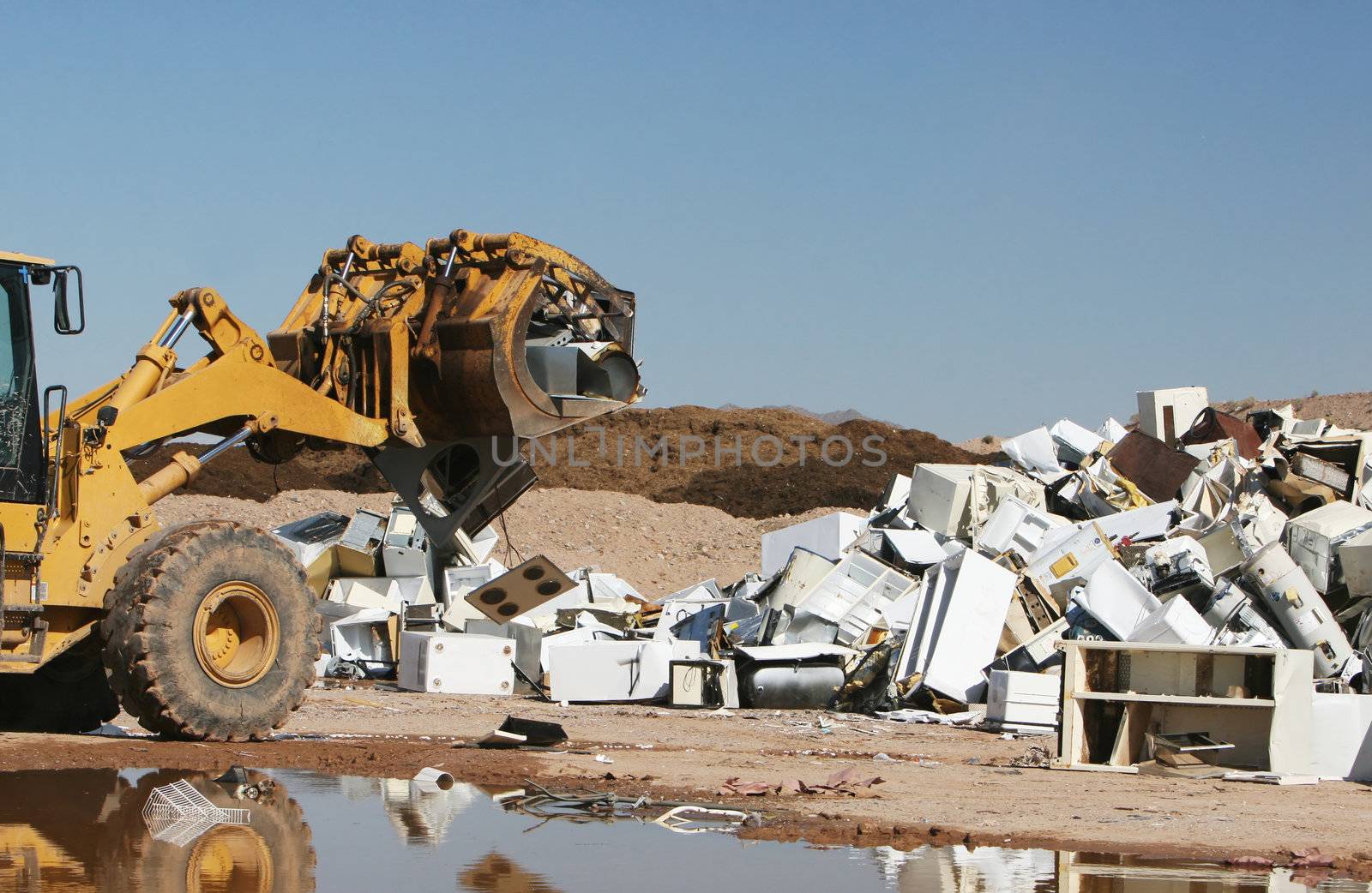 A bulldozer crushes and moves a giant stack of refrigerators, air conditioners, and stoves.