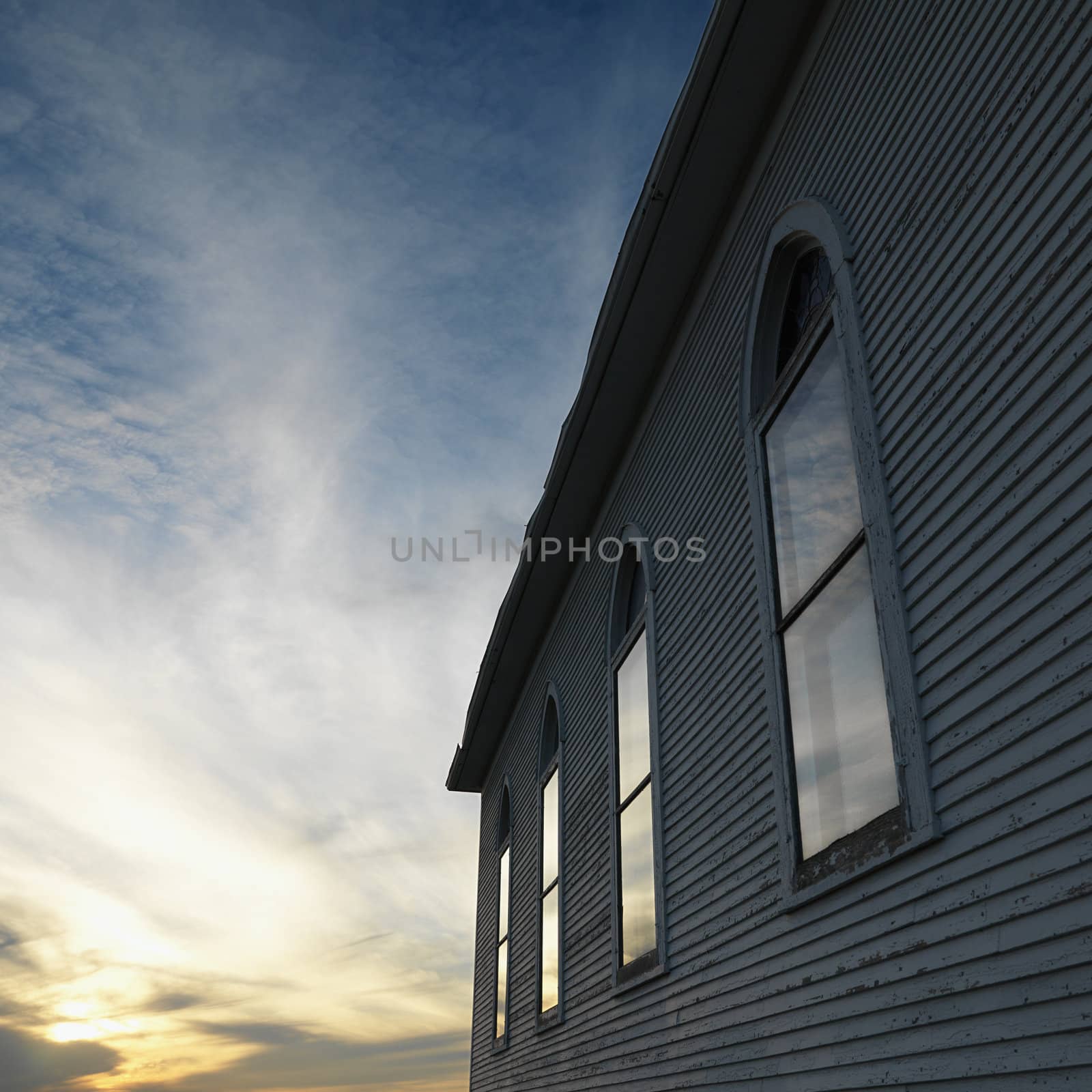 Sunset and clouds behind side view of building with arched windows and wooden siding.