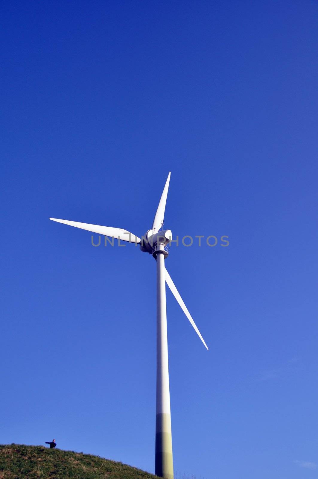 Wind turbine as alternative energy source on green hill with boy pointing to the left and shiny blue sky in the background
