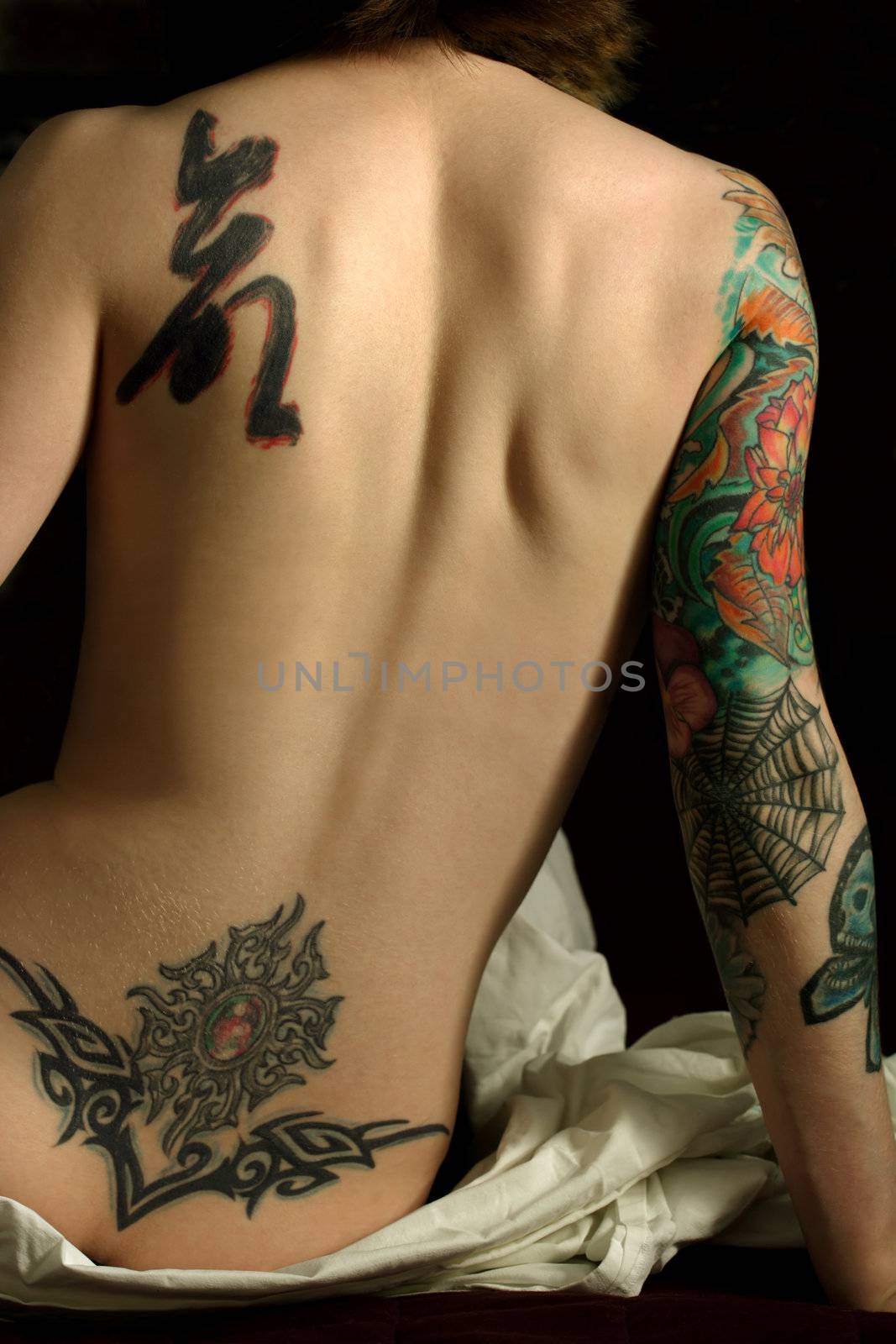 A young slim women with arm and back tattoos sitting on the bed in a dark room.
