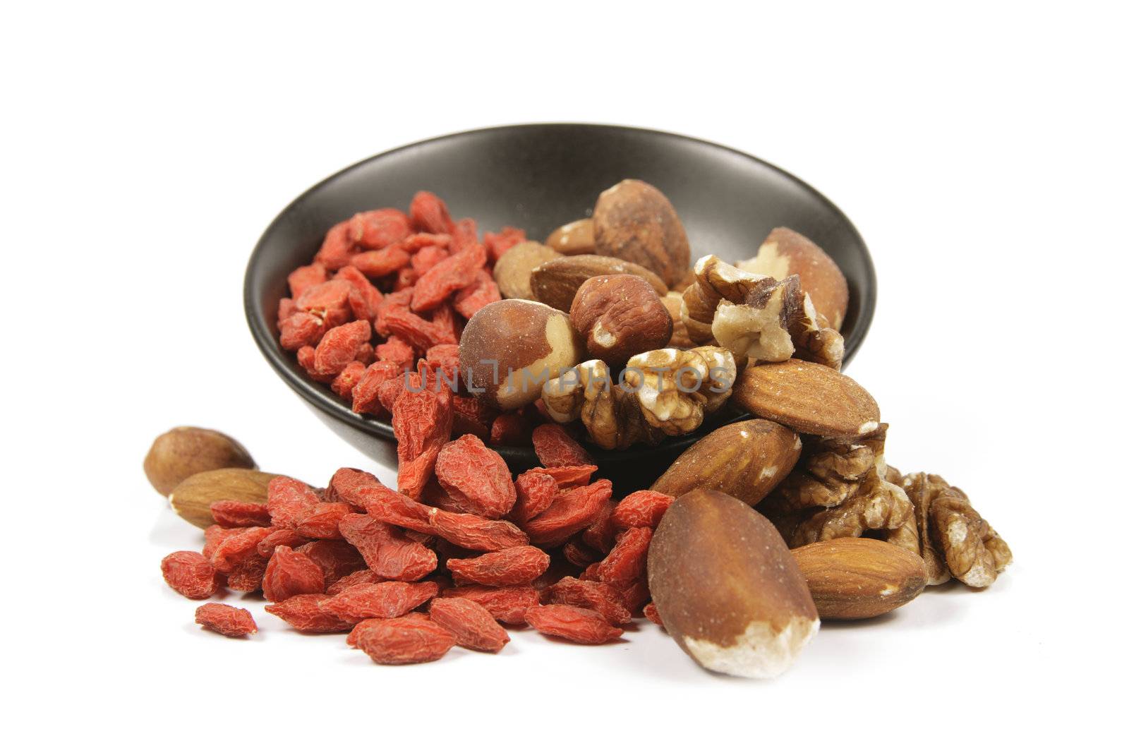 Red dry goji berries with mixed nuts spilling from a small black bowl on a reflective white background