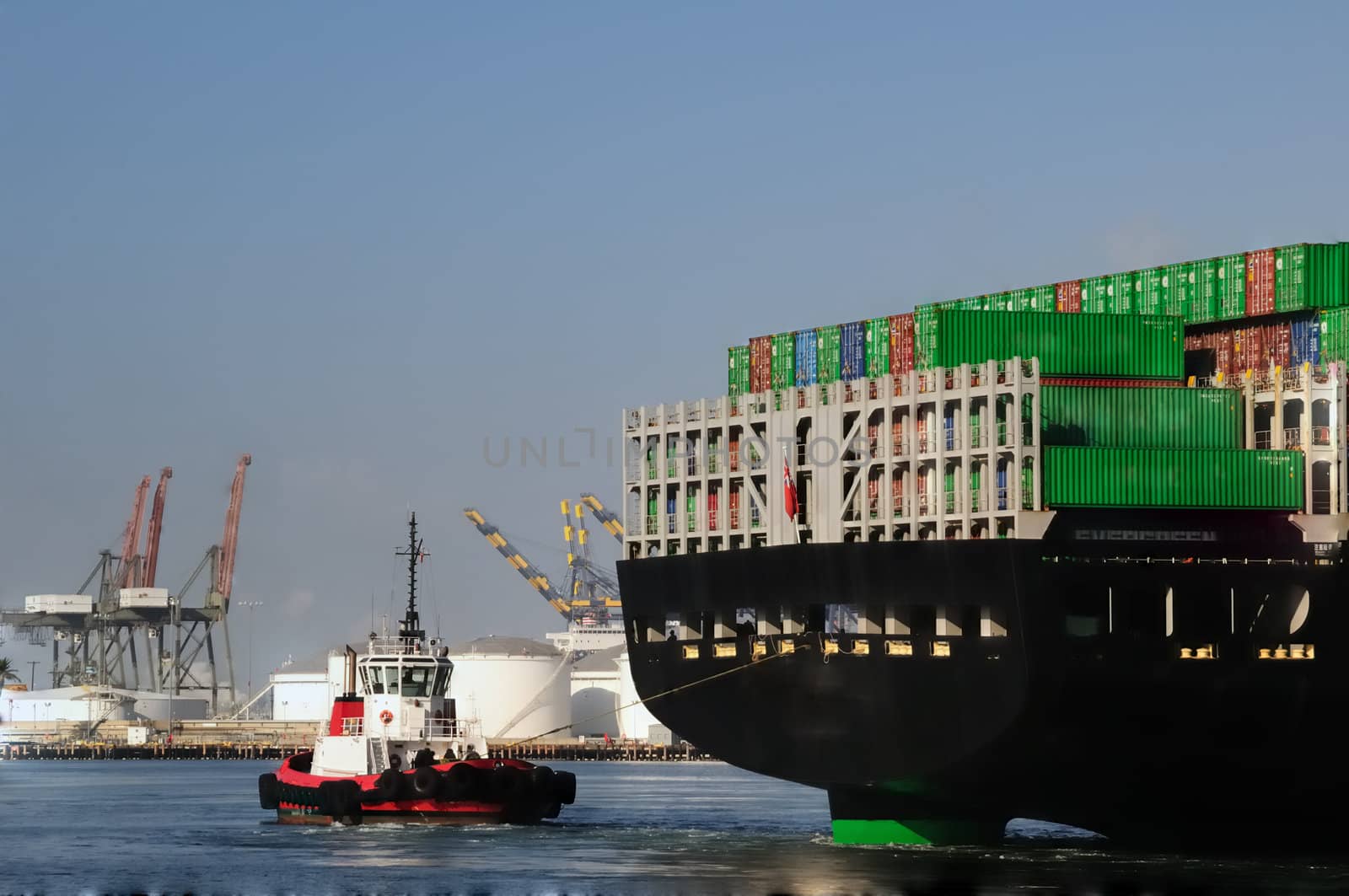 View from behind as a red tugboat pulls a massive merchant vessel around in a narrow harbor channel, with loading cranes in the background.