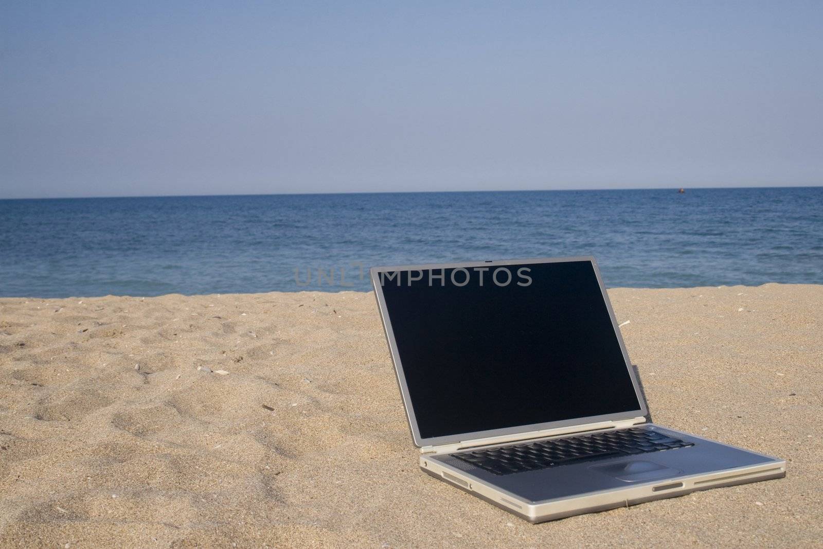 Laptop sitting on sand with sea and blue sky behind