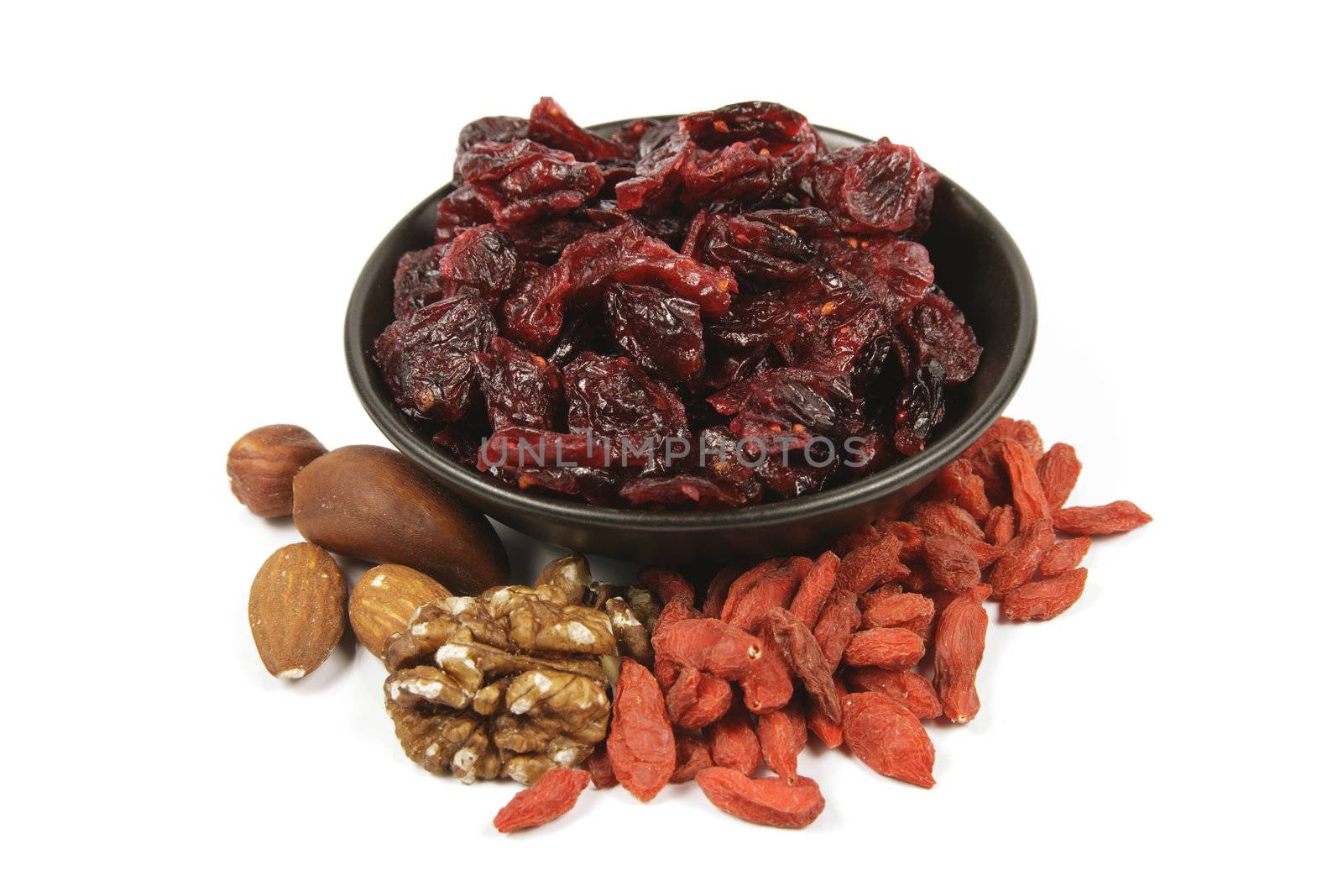 Red ripe dried cranberries in a small black bowl with mixed nuts and goji berries on a reflective white background
