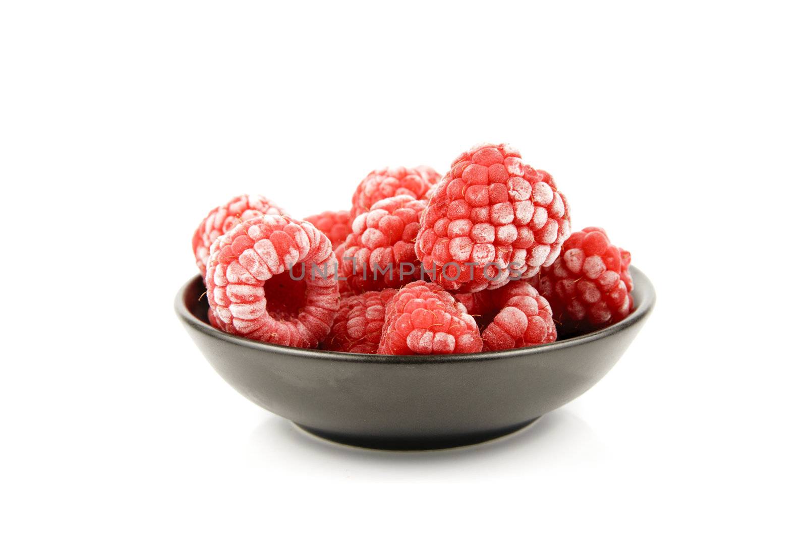 Red ripe frozen raspberries in a small black bowl on a reflective white background