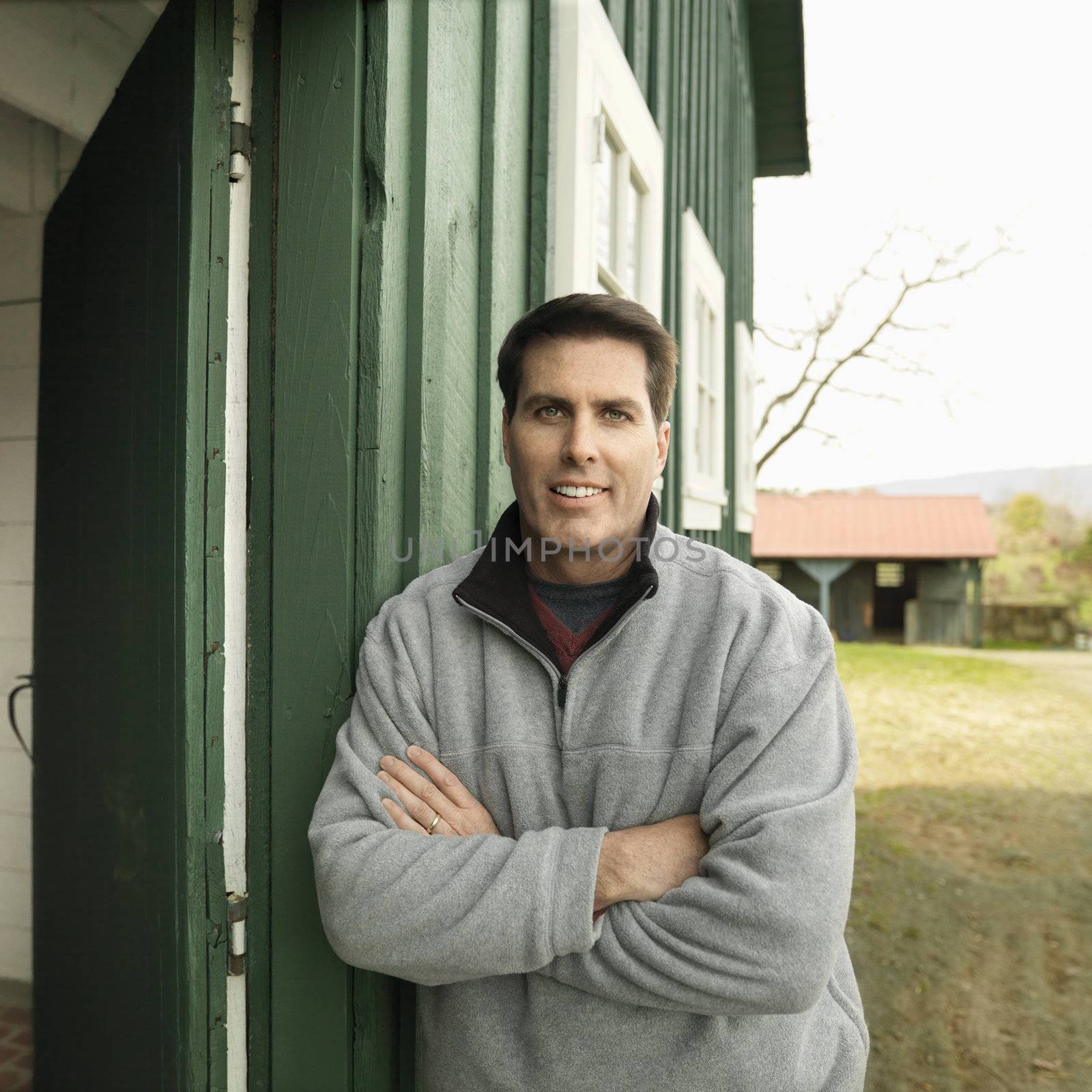 Caucasian man leaning against building smiling in rural setting.