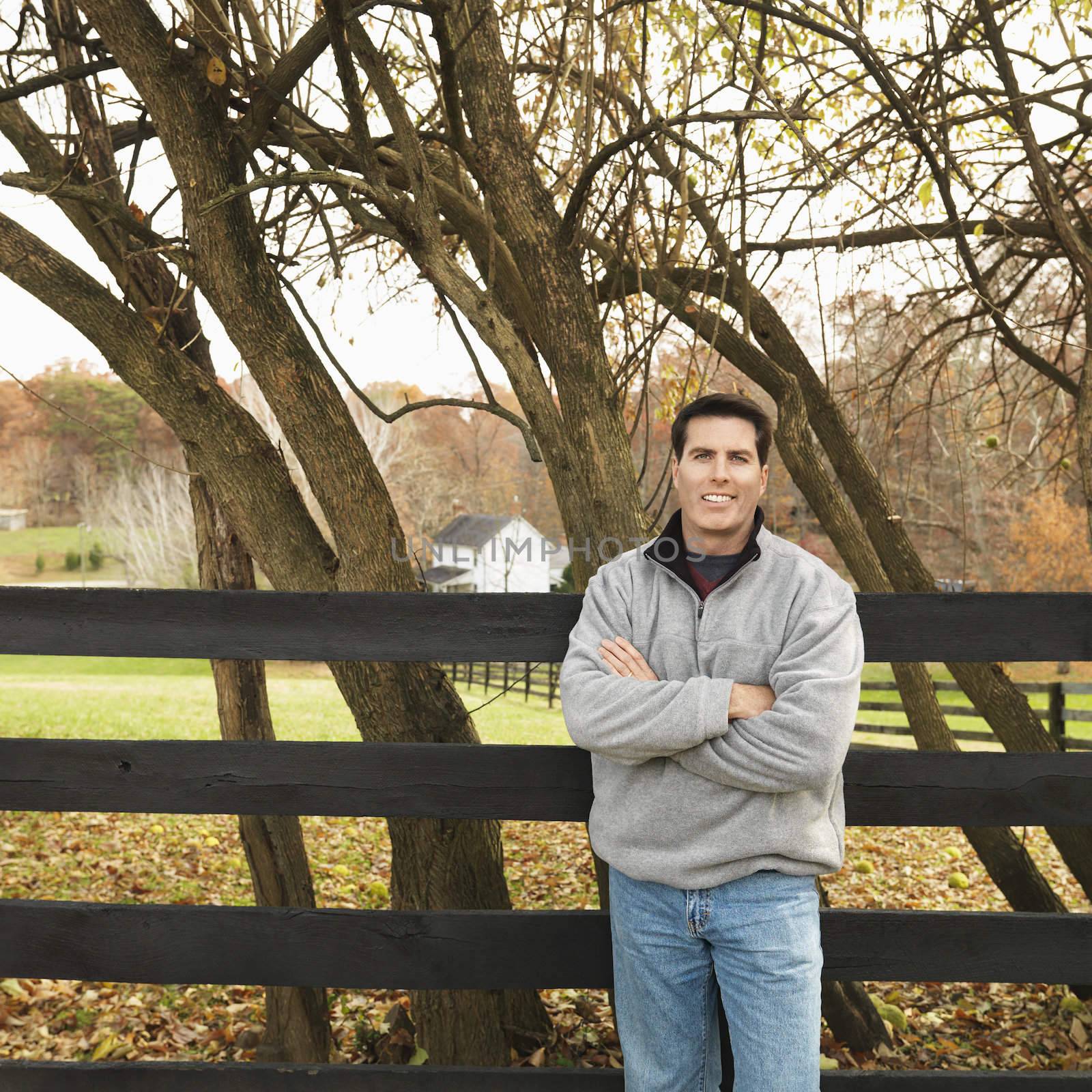 Caucasian man leaning against fence smiling in rural setting.