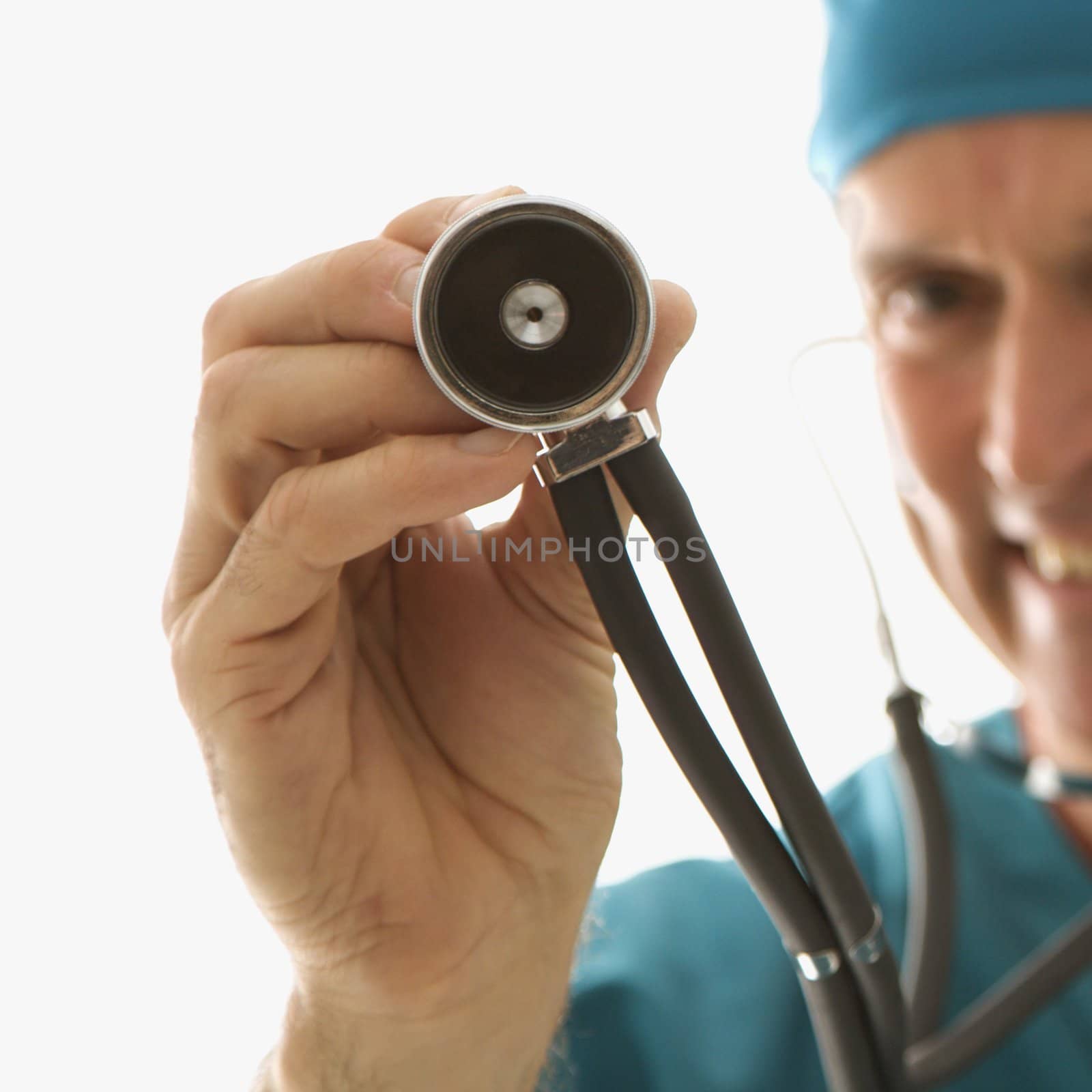 Mid-adult Caucasian male doctor in scrubs holding a stethoscope out towards viewer.