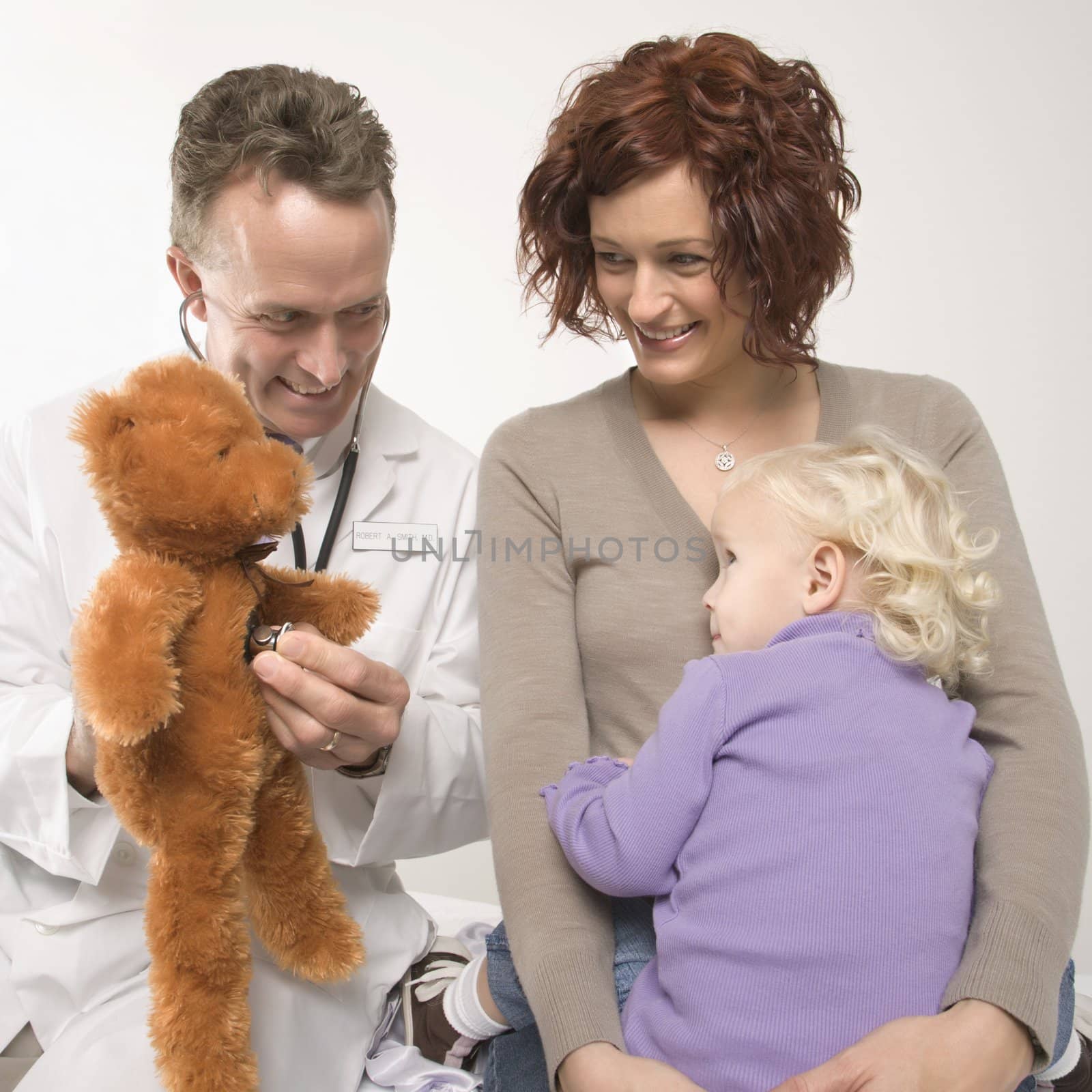 Middle-aged adult Caucasian male doctor holding stethoscope to teddy bear while Caucasian mother and daughter watch.