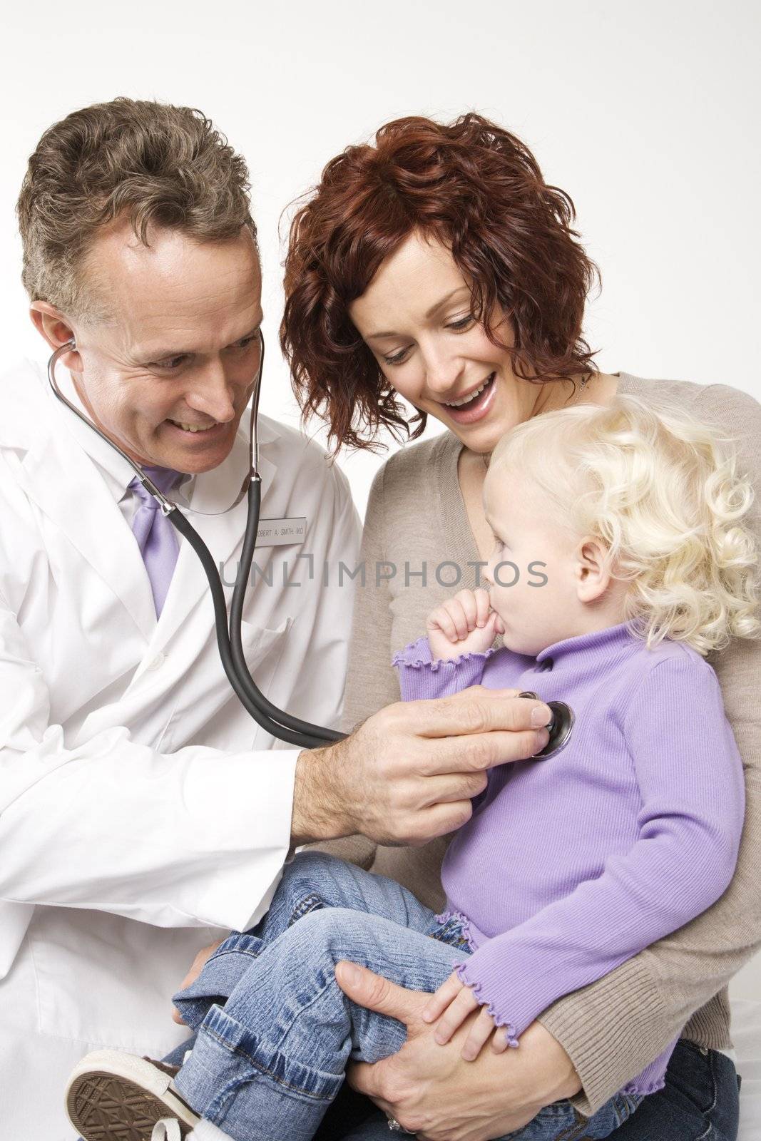 Middle-aged adult Caucasian male doctor holding stethoscope to female toddler's chest with mother watching.