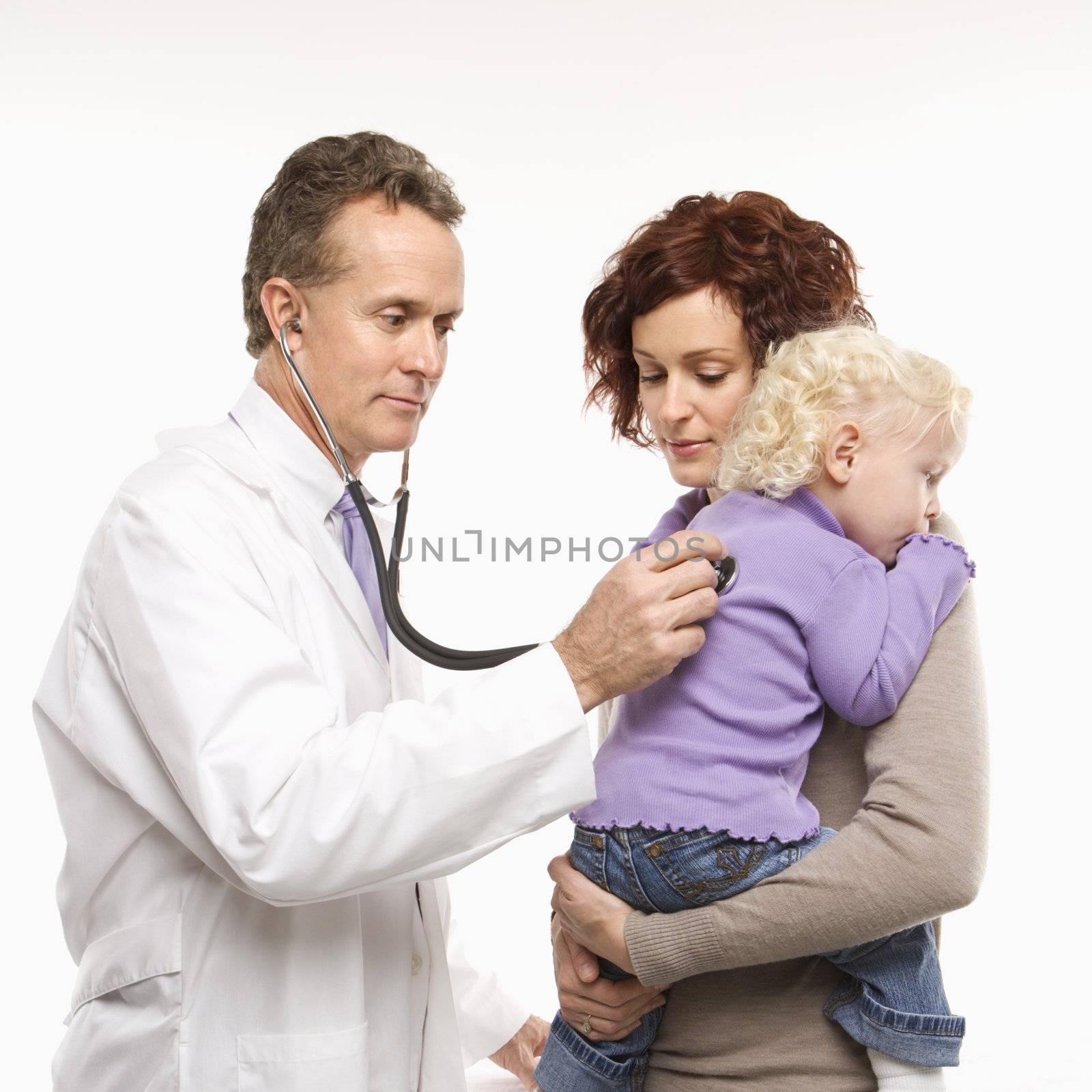 Middle-aged adult Caucasian male doctor holding stethoscope to female toddler's back with mother watching.