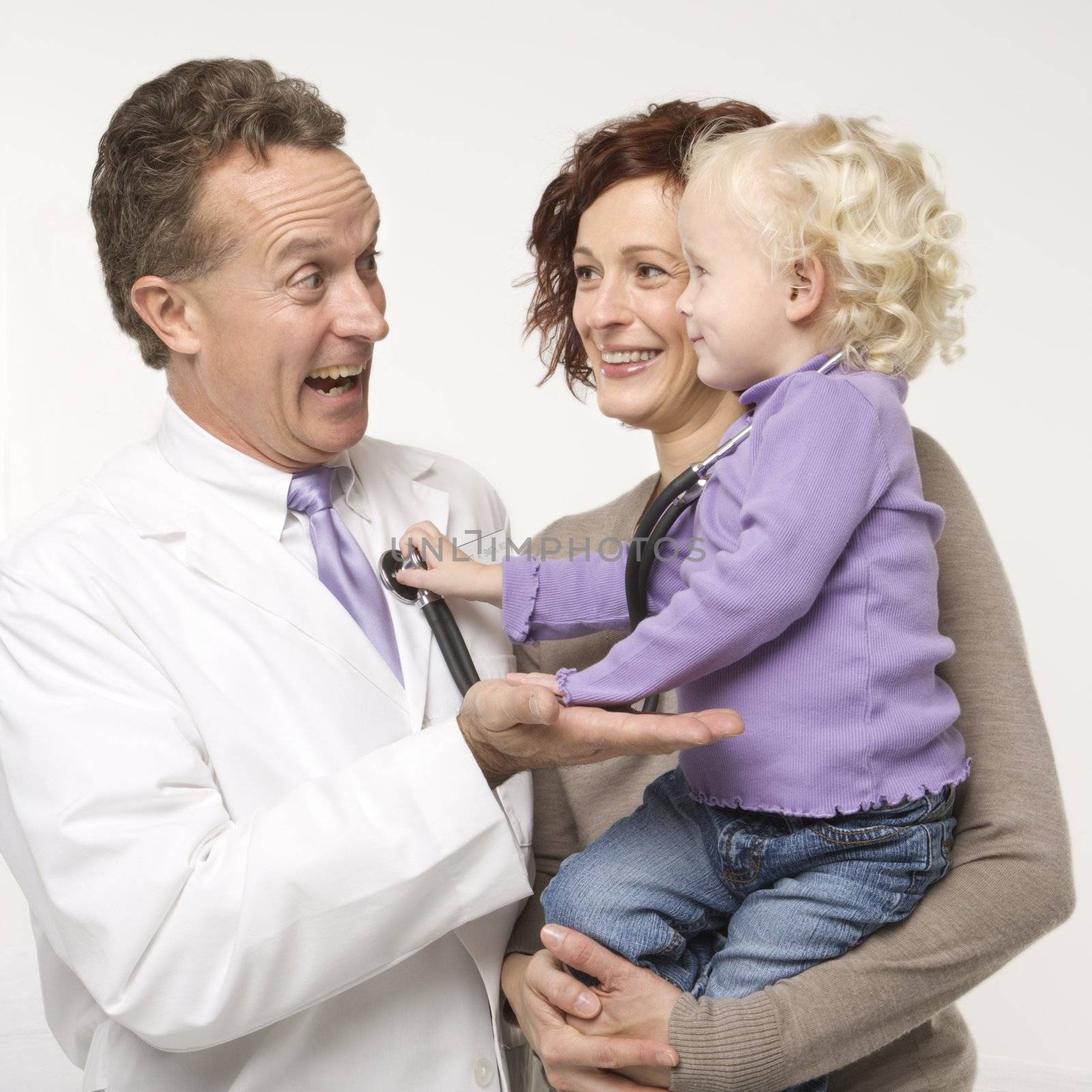 Caucasian female toddler holding stethoscope up to middle-aged male doctor smiling.