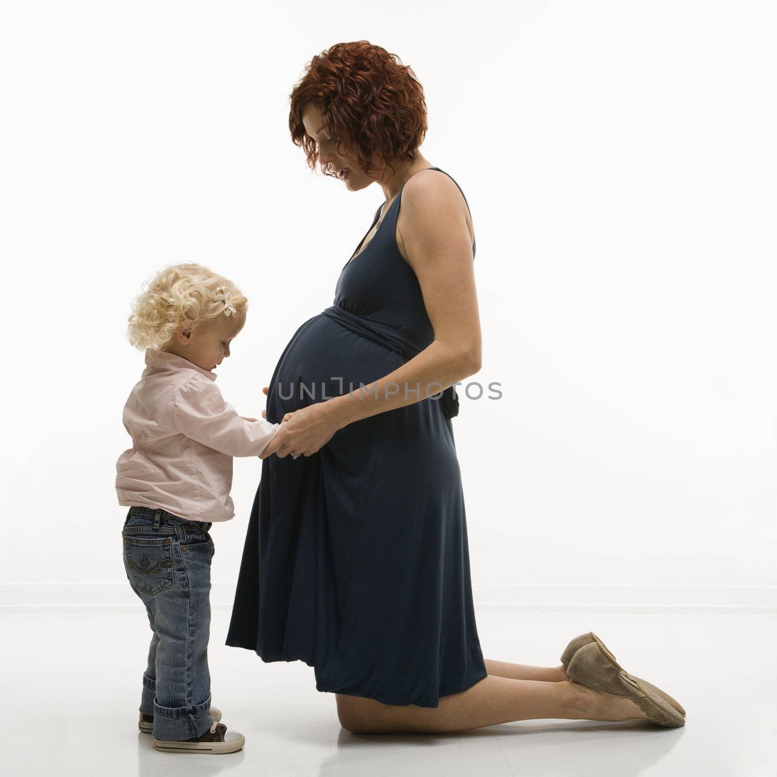 Caucasion mid-adult attractive pregnant woman kneeling in front of female toddler who is touching and looking at her belly.