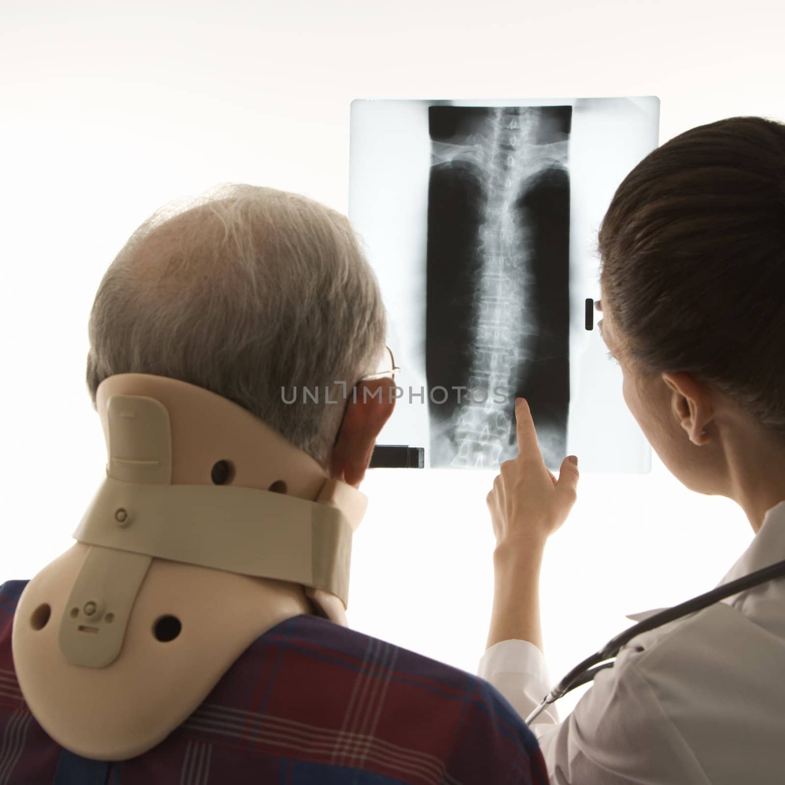 Over the shoulders view of mid-adult Caucasian female pointing at an x-ray as elderly Caucasian male in neck brace looks on.
