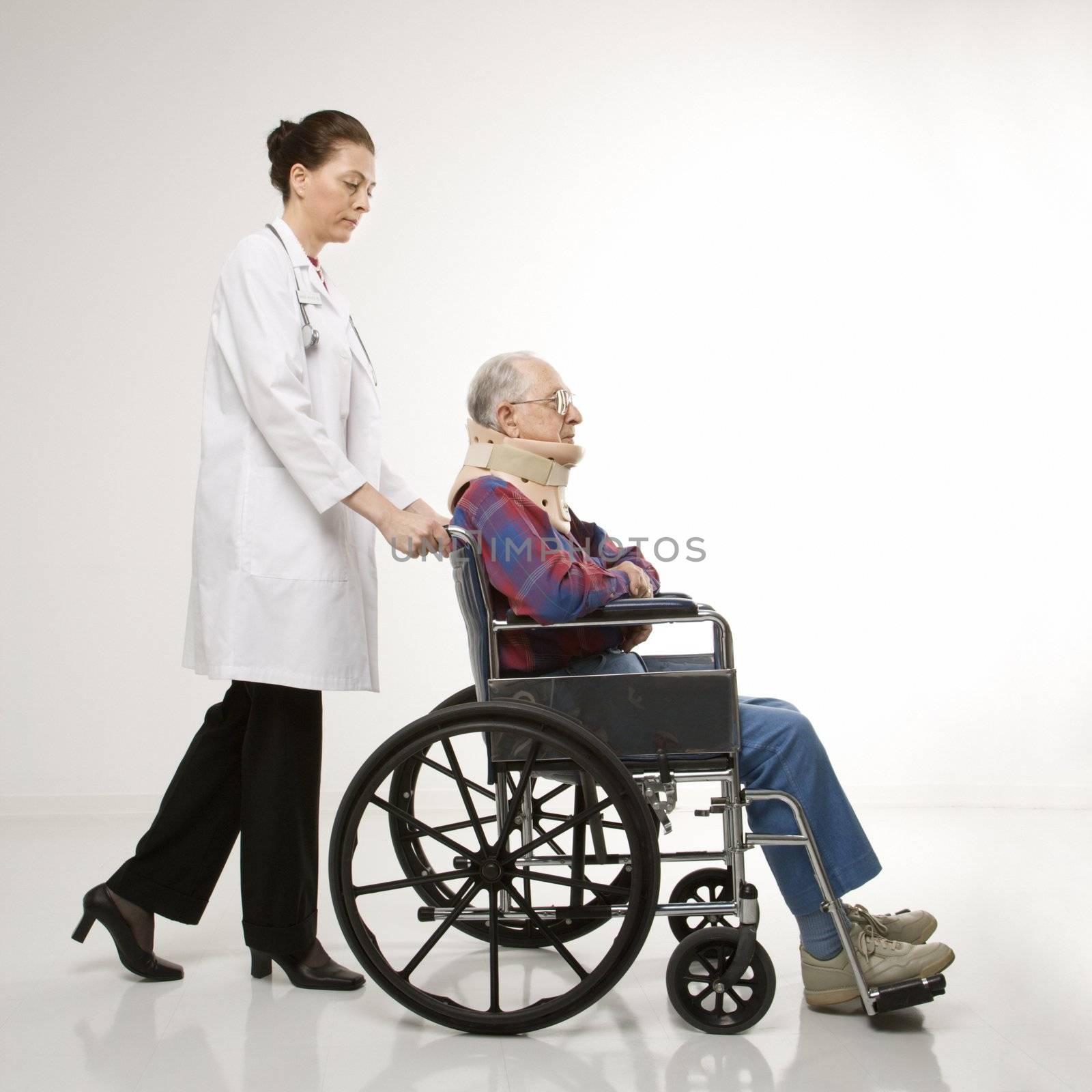 Mid-adult Caucasian female doctor pushing elderly Caucasian male with neck brace in wheelchair.