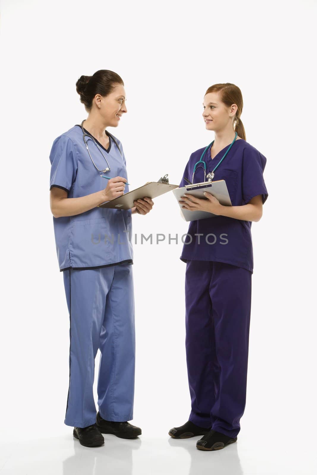 Portrait of Caucasian women doctors in medical scrubs standing talking holding medical charts against white background.