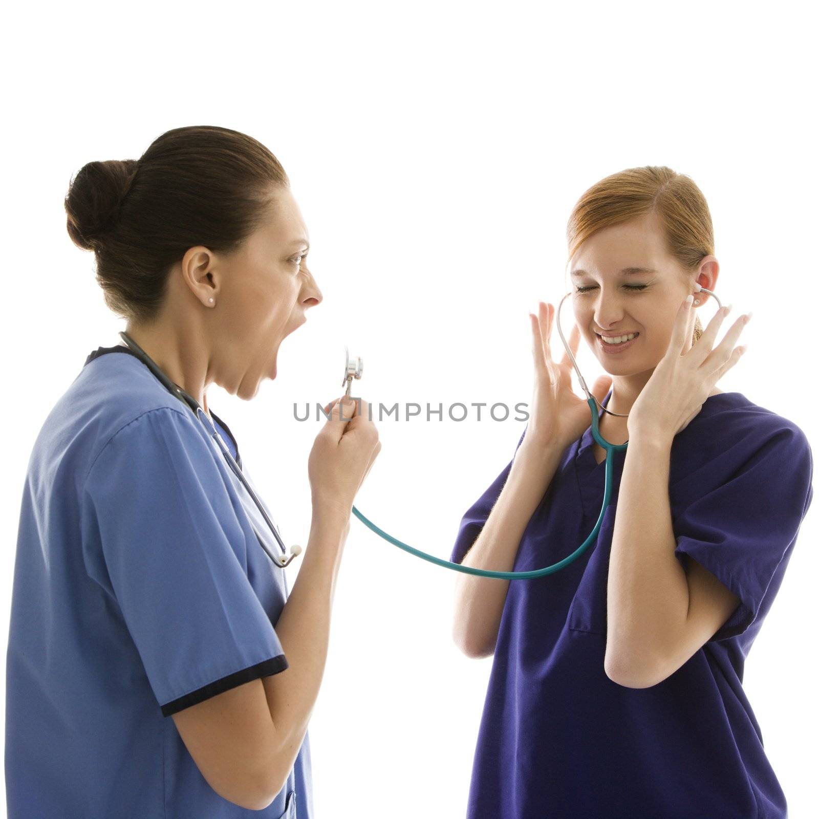 Portrait of Caucasian healthcare workers wearing scrubs with one yelling into stethoscope that is attached to other's ears against white background.
