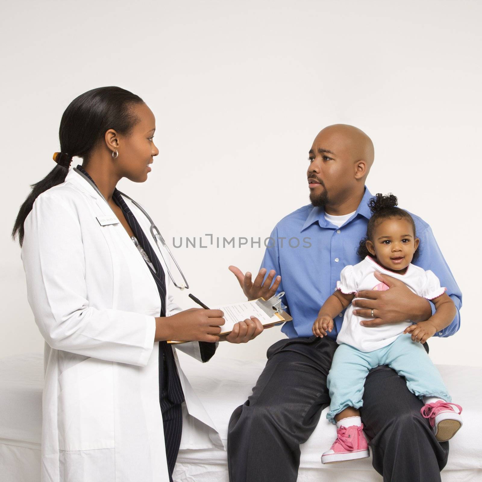 African-American father holding baby girl talking to female pediatrician.