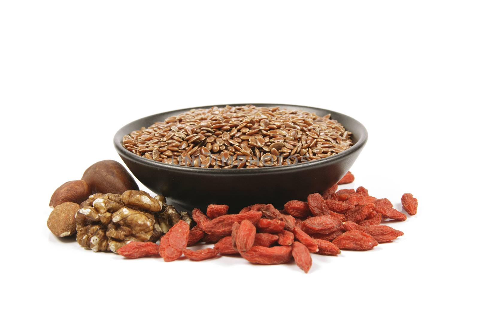 Brown linseed seeds in a small black bowl with mixed nuts and goji berries on a reflective white background