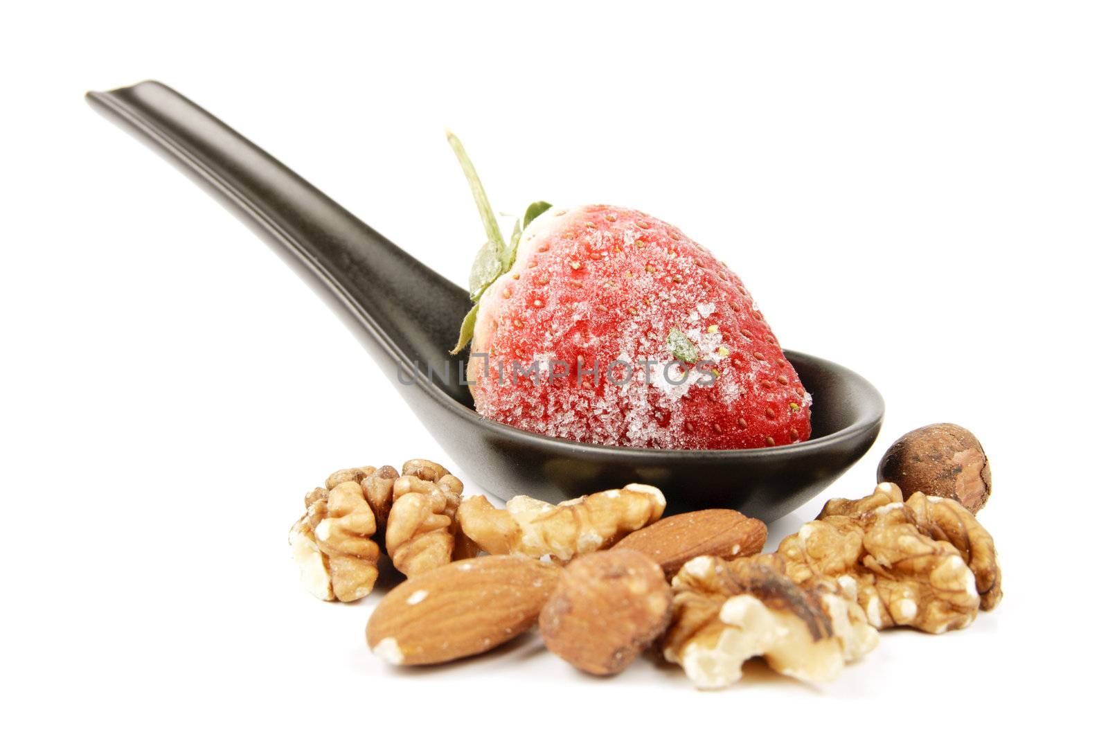 Red ripe frozen strawberry on a small black spoon with mixed nuts on a reflective white background