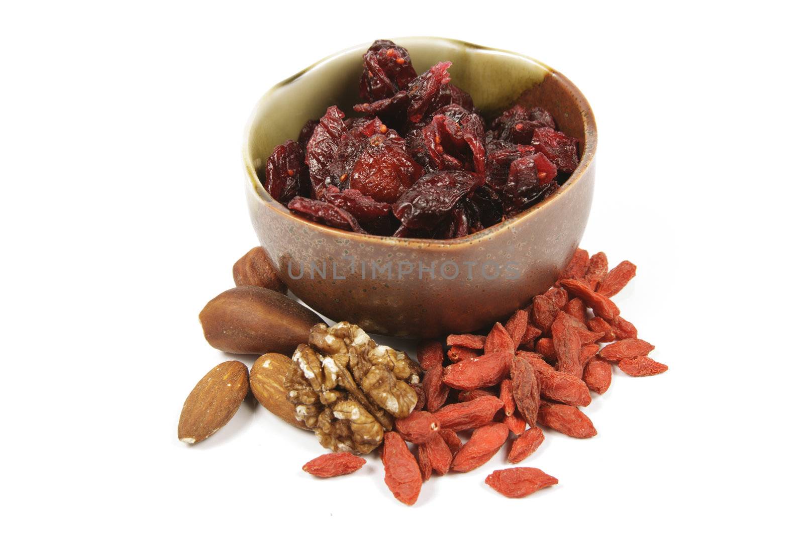 Red ripe dried cranberries in a small green and brown bowl with mixed nuts and goji berries on a reflective white background
