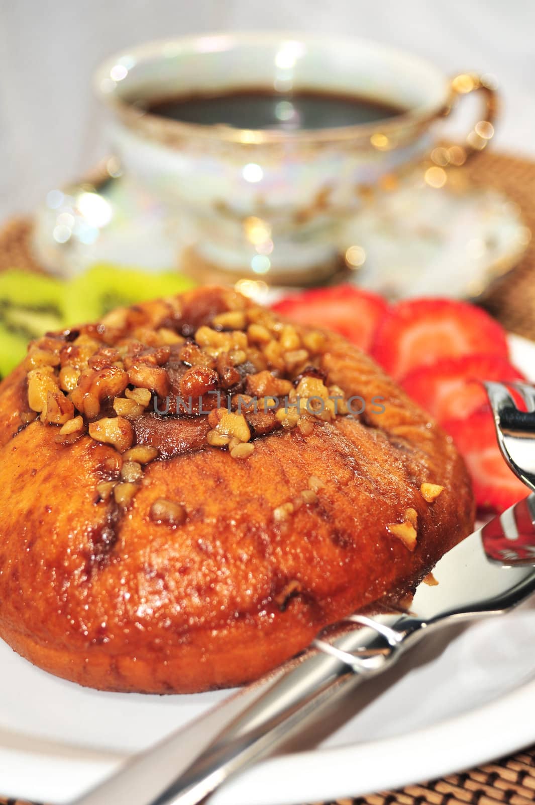 A fresh Cinnamon Roll topped with Walnuts, sliced Strawberries and Kiwi on the side and a hot cup of coffee.