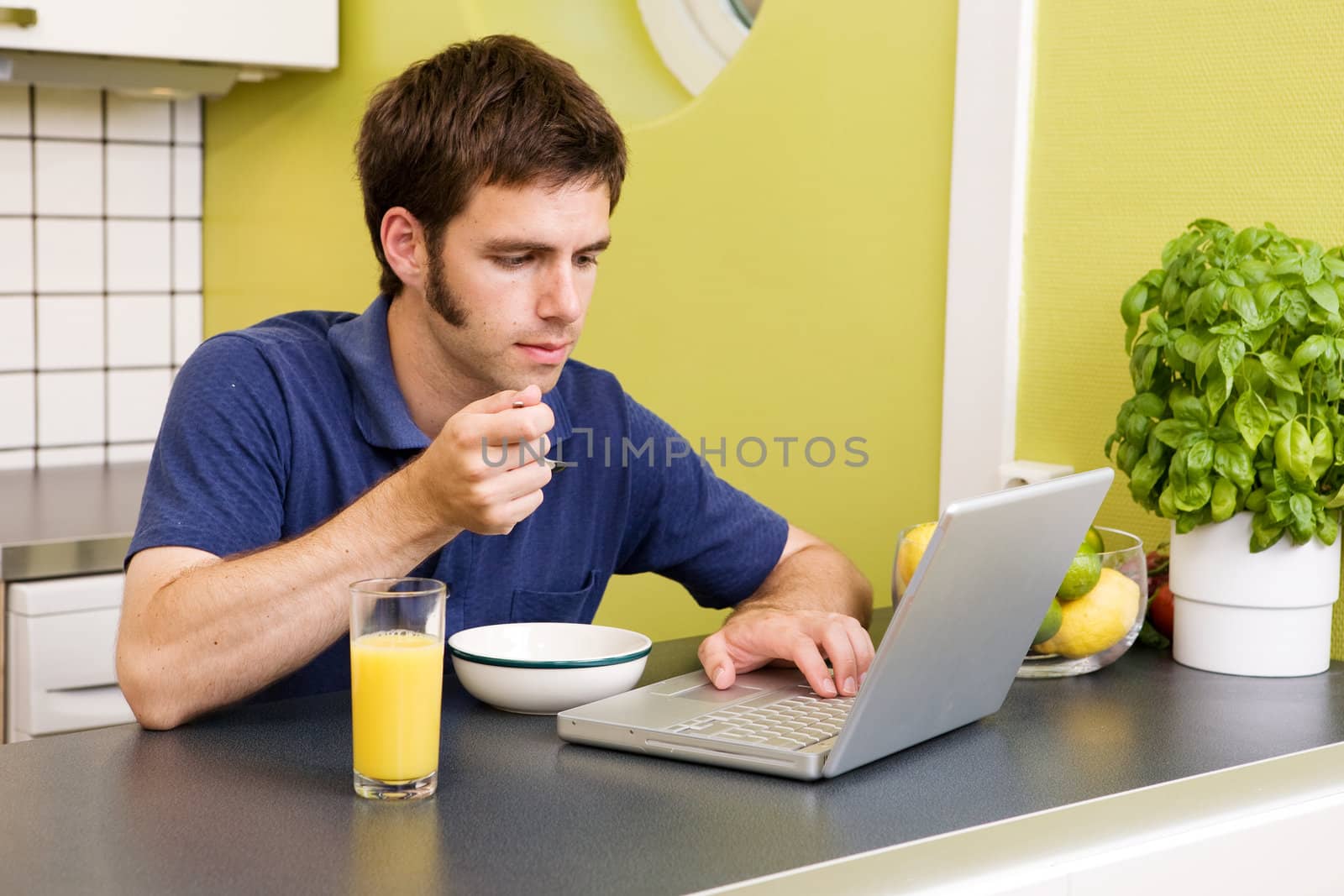 A young male works on the computer in the kicthen while eating breakfast