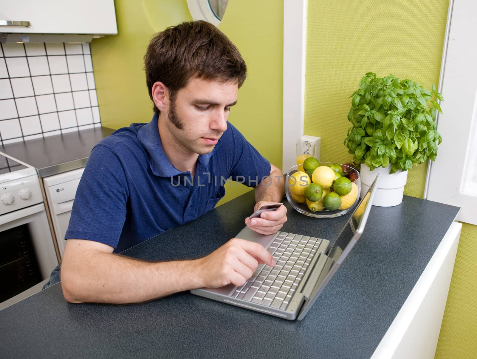 A young male shops online with a credit card at home in the kitchen.