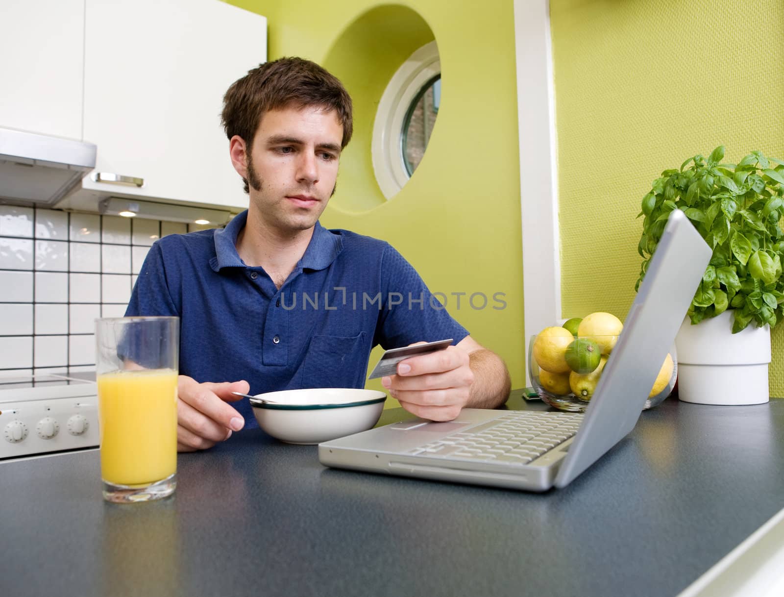 A young male shops online with a credit card in his kitchen while eating breakfast