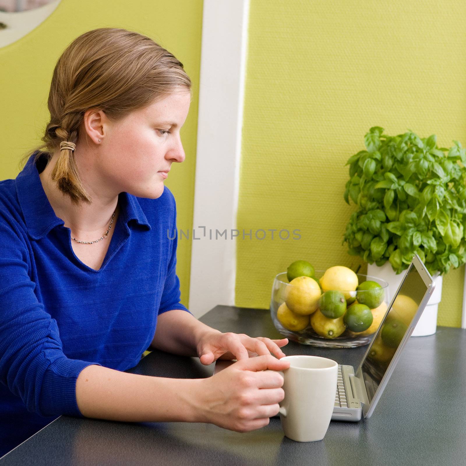 A young woman sitting in the kitchen with a coffee and a computer looking at the camera.