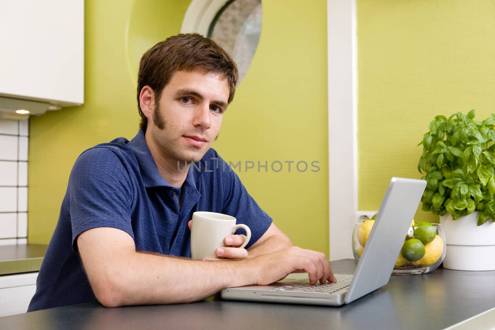 A young man uses the computer in the kitchen while enjoying a warm drink