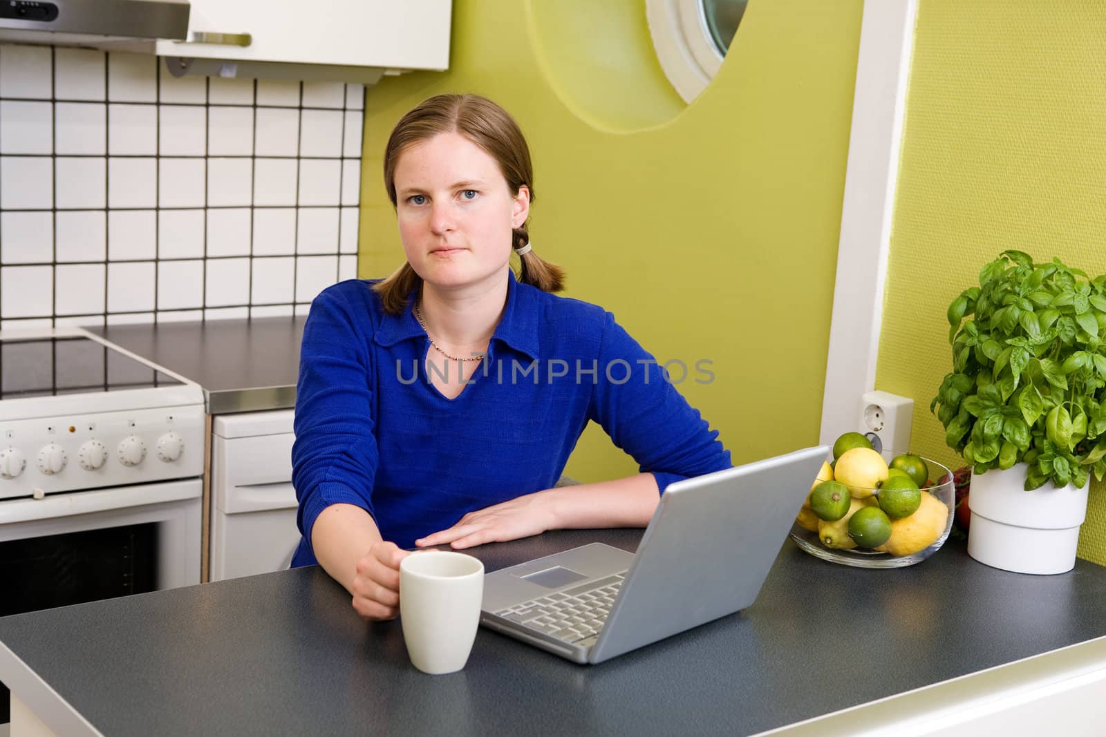 A young woman sitting in the kitchen with a coffee and a computer looking at the camera.