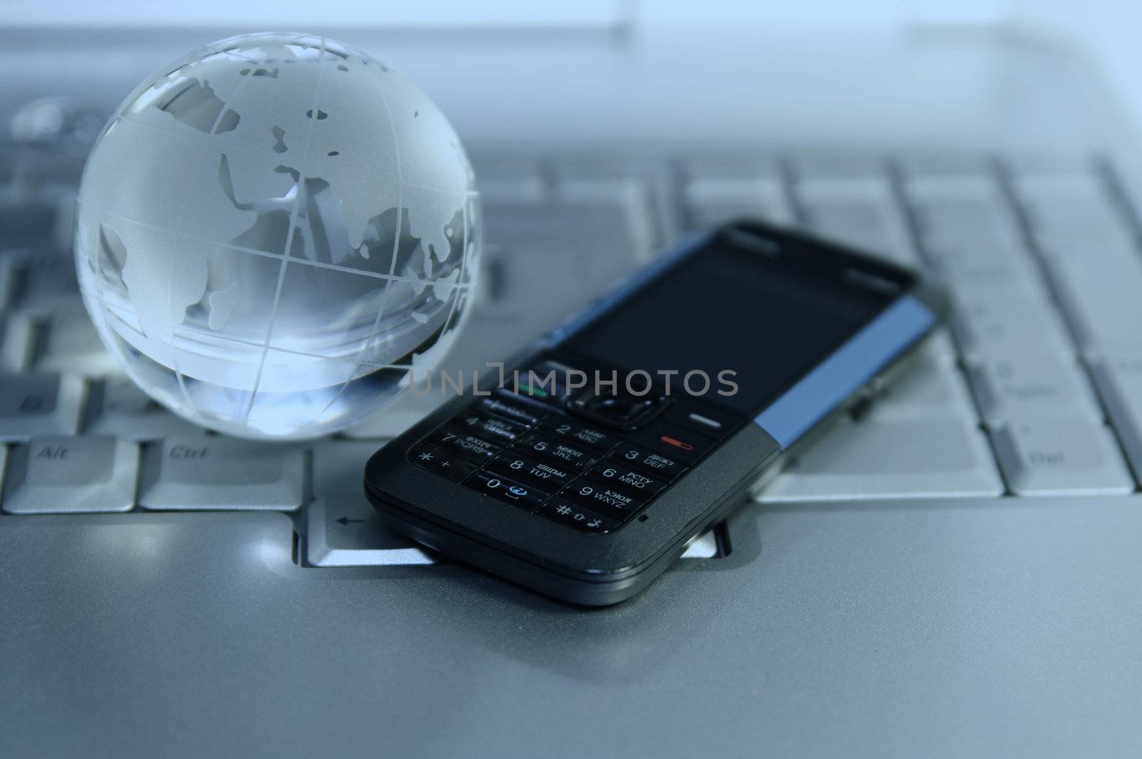 Blue tinted image of cellular and glass globe on laptop keyboard. Narrow DOF. Focus on mobile phone buttons