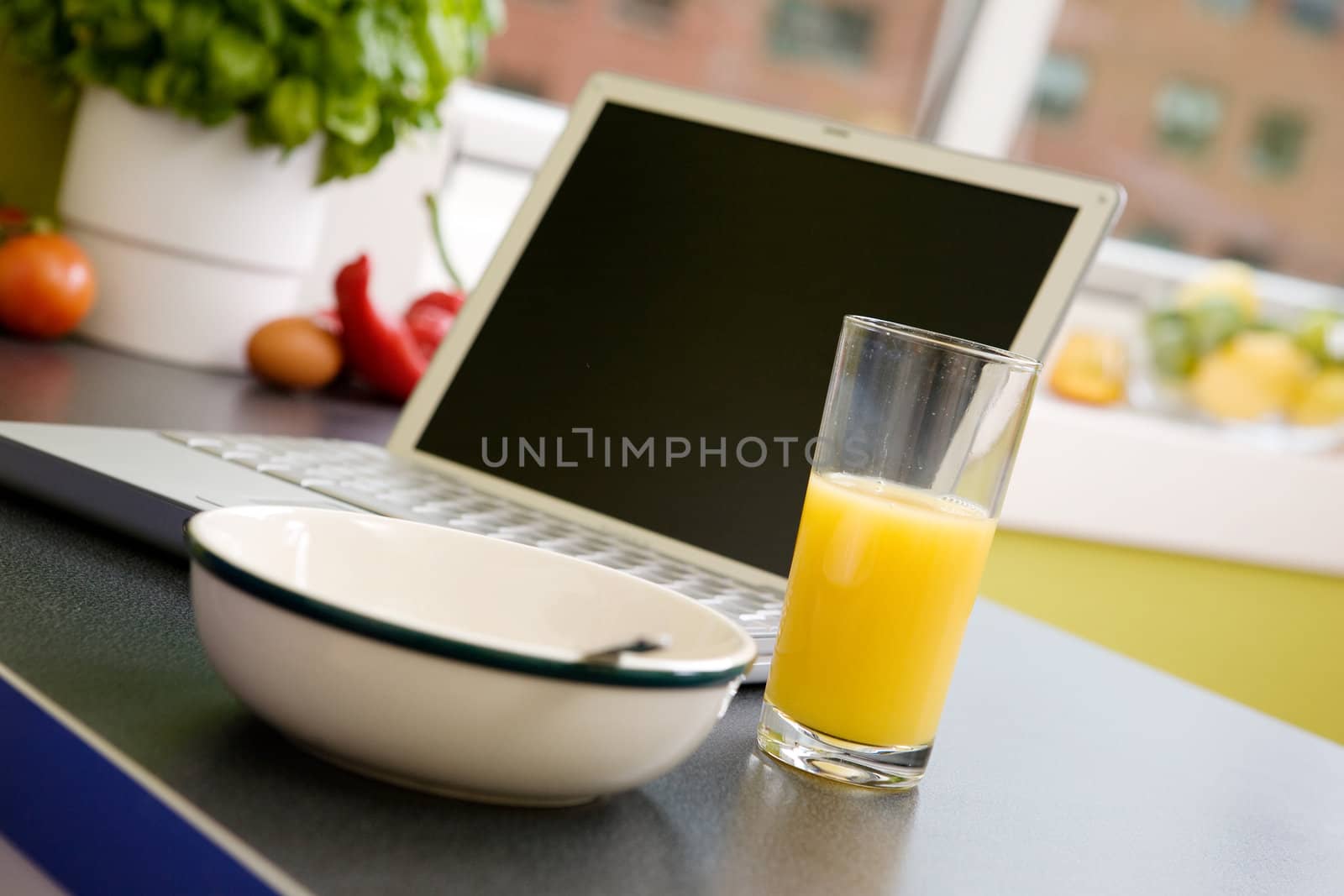 A computer in the kitchen with a bowl of soupr or cereal and orange juice - shallow depth of field with focus on juice