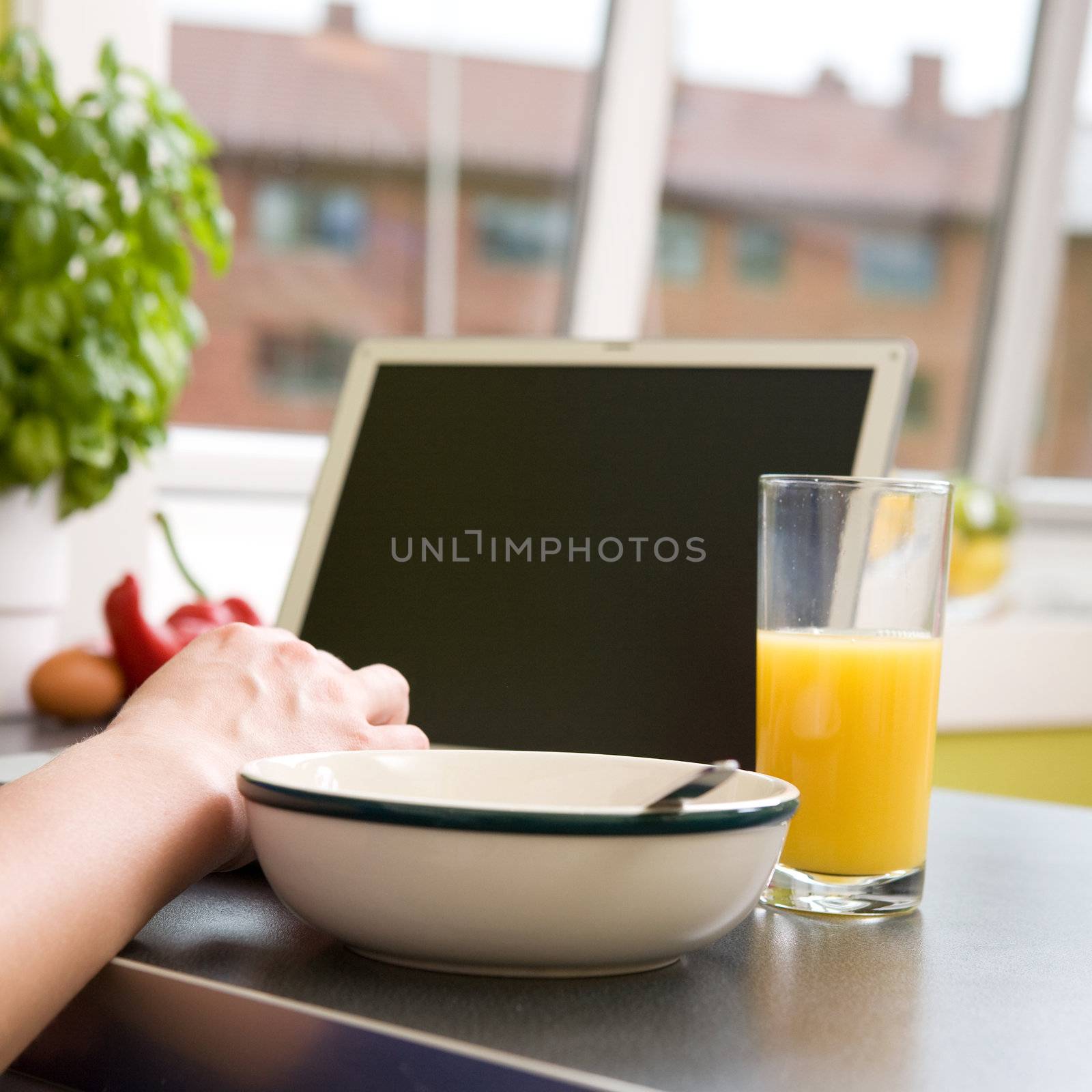 A computer in the kitchen with a bowl of soupr or cereal and orange juice - shallow depth of field with focus on juice and hand