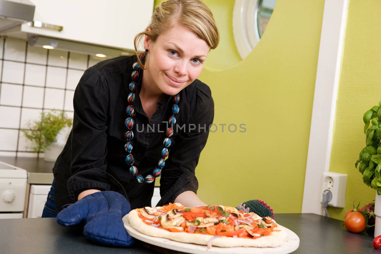 A young woman smiles at the camera with a fresh homemade pizza out of the oven in her apartment kitchen.