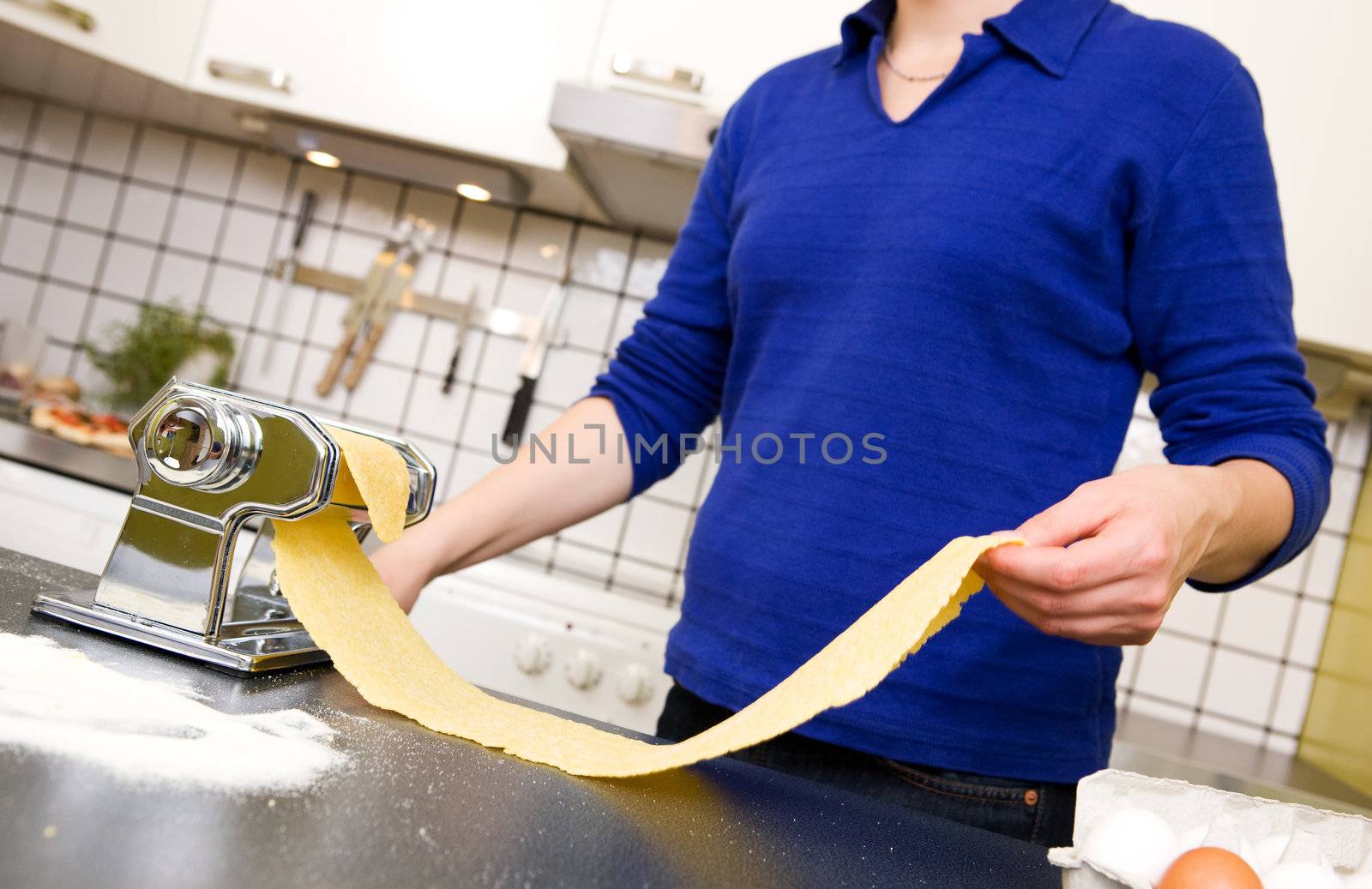 A young female stretching out pasta over the counter from a manual pasta machine at home in the kitchen.