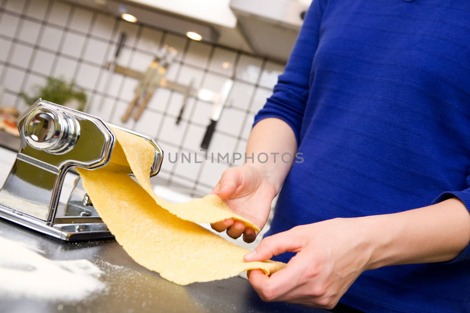 A young female stretching out pasta over the counter from a manual pasta machine at home in the kitchen.