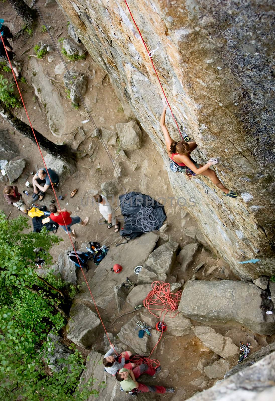 An eager female climber on a steep rock face looks for the next hold - viewed from above.  Shallow depth of field is used to isolated the climber.  A belayer and other climbers look on in anticipation.