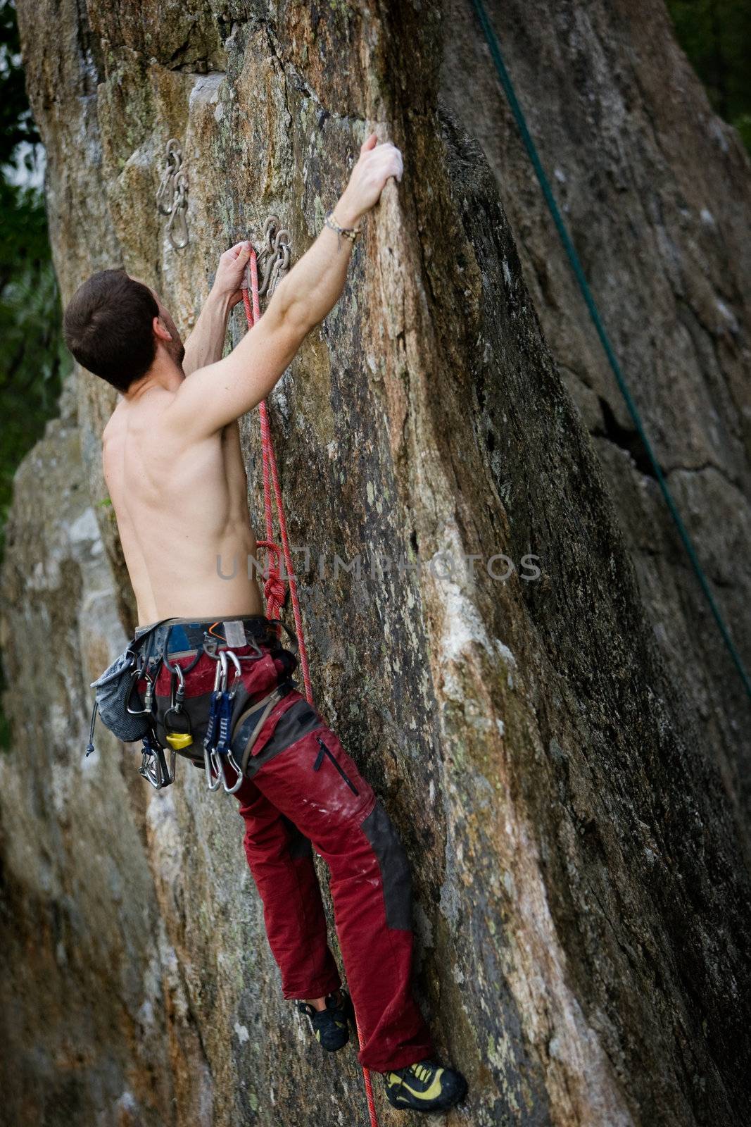 A male climber, viewed from above, climbs a very high and steep crag.  The image is taken as the climber clips into the bolt.