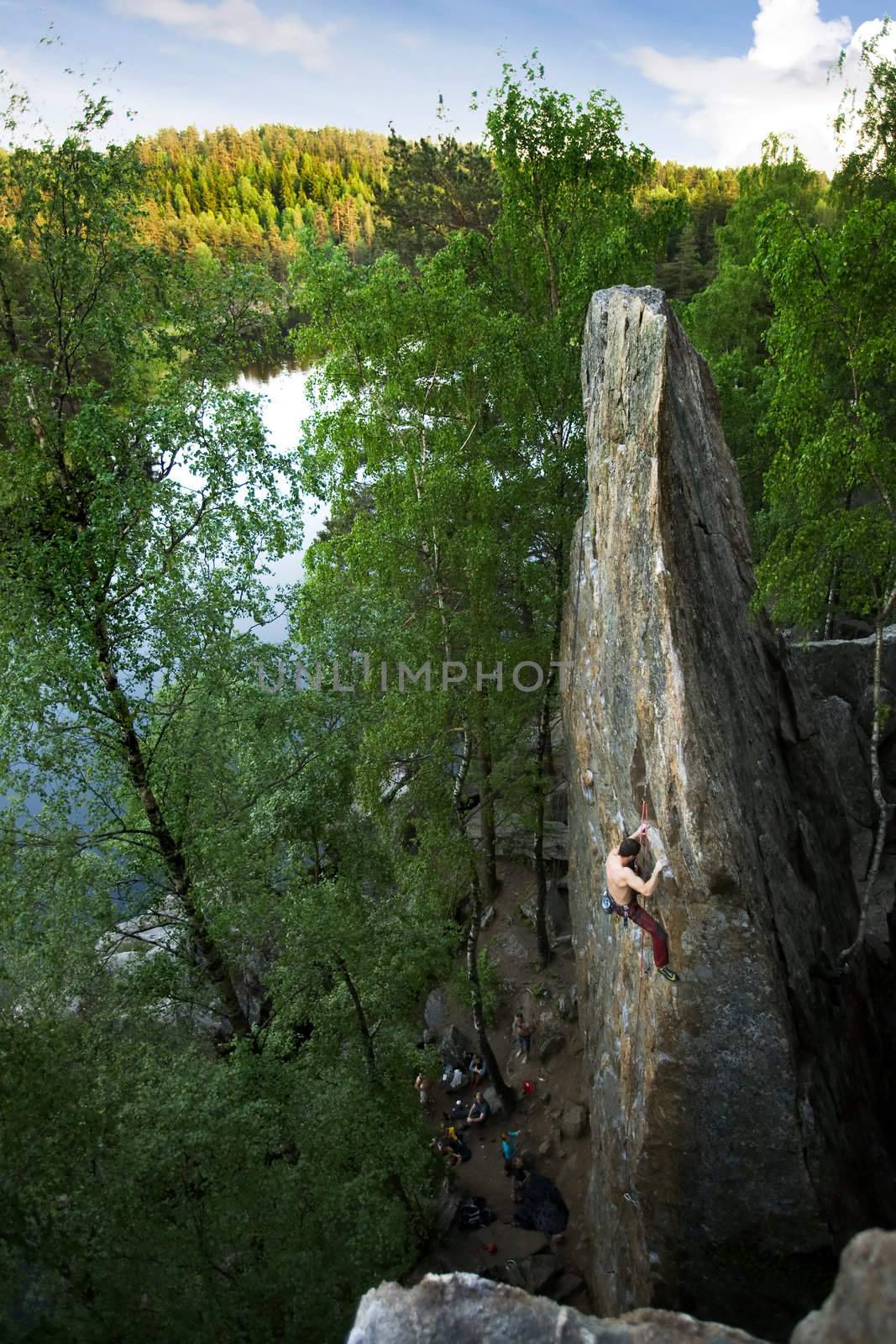 A male climber, viewed from above, climbs a very high and steep crag. A beautiful lake and landscape create a breathtaking backdrop.