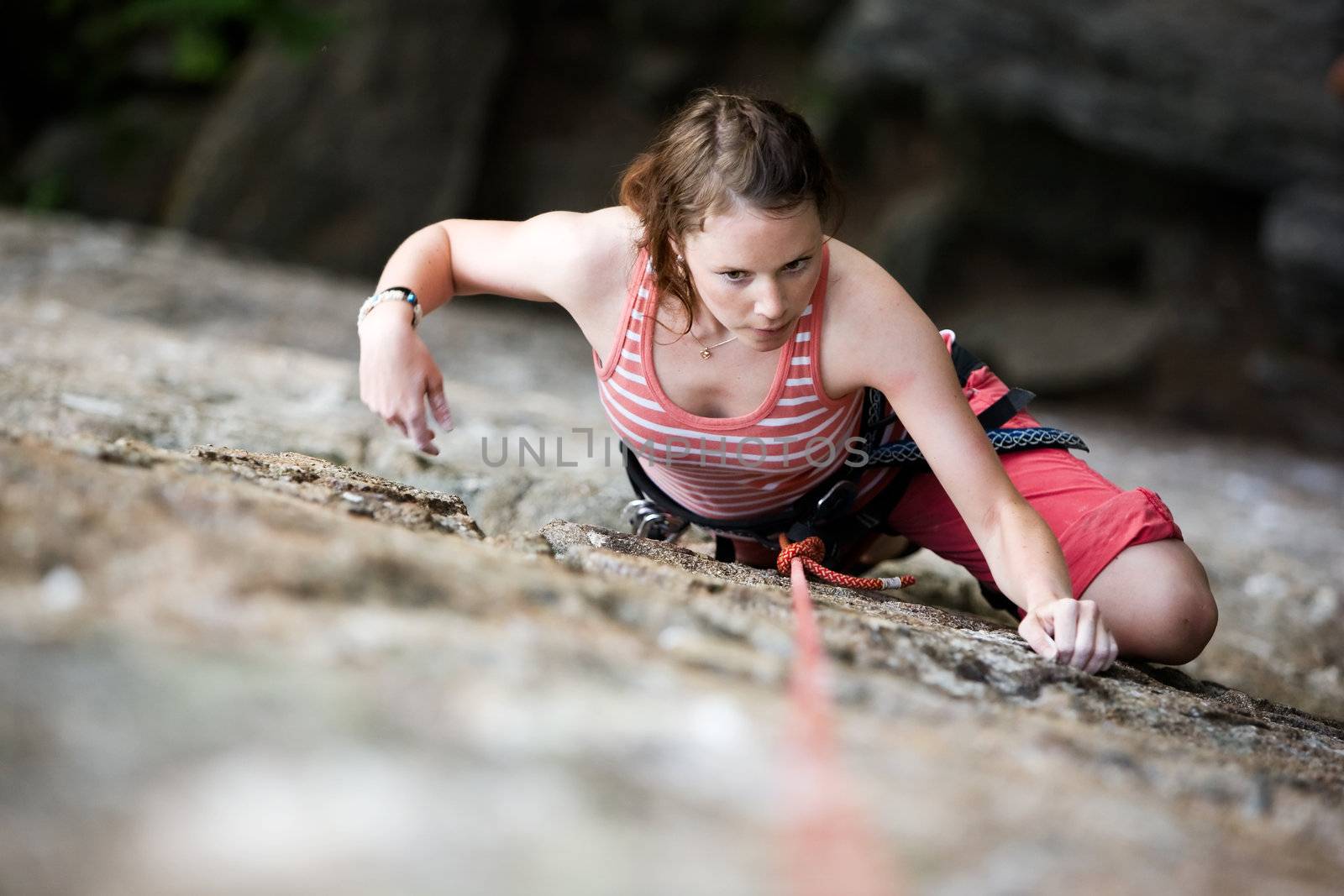 A female climber on a steep rock face viewed from above with the belayer in the background.  Shallow depth of field is used to isolated the climber.