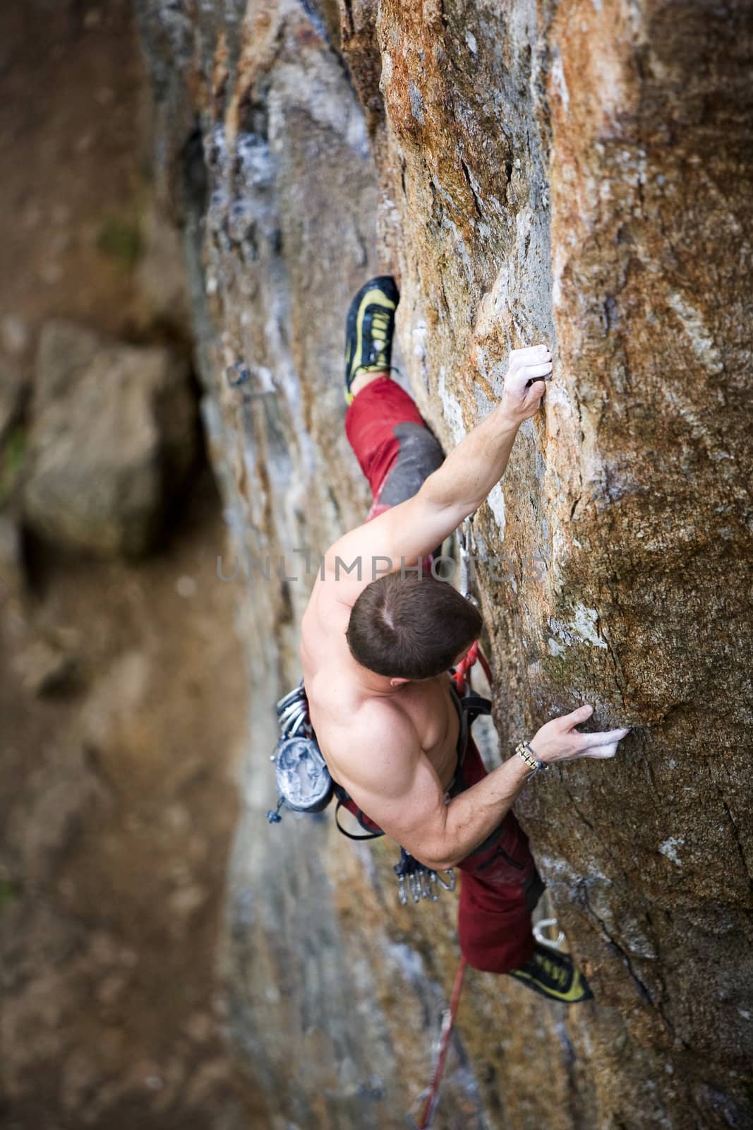 A male climber viewed from above climbs a steep crag. Shallow depth of field is used to isolate the climber with focus on the hands and head