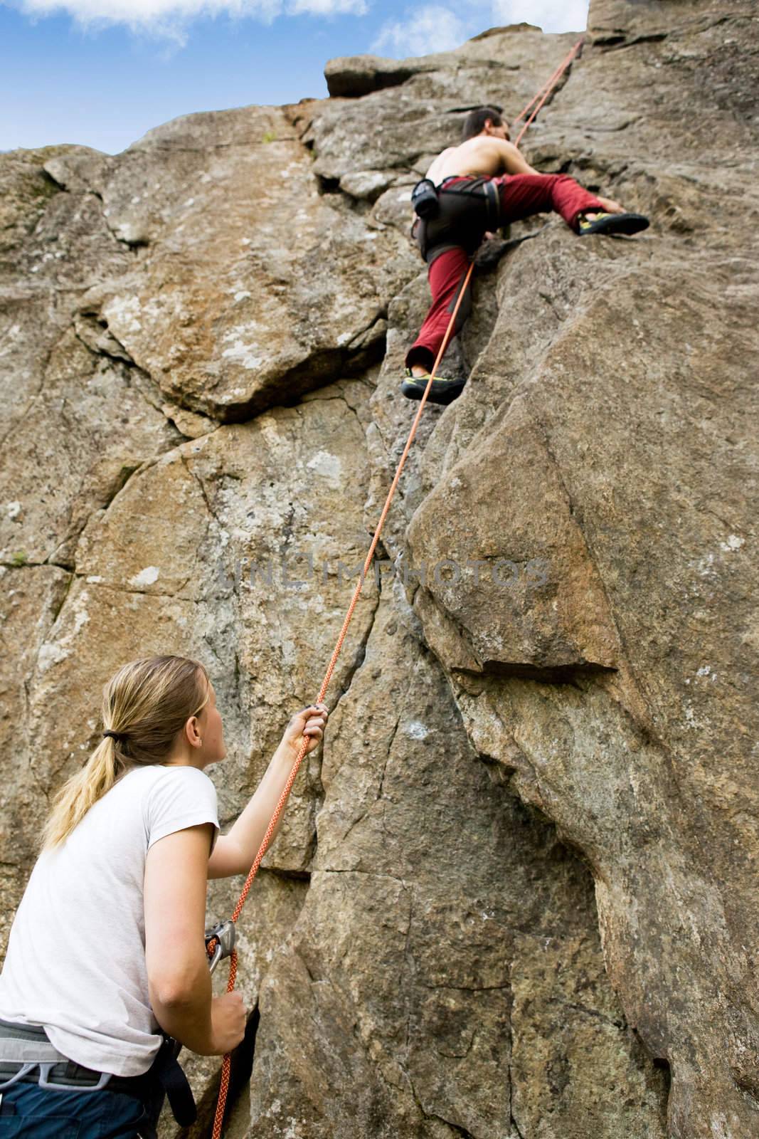 A female belaying a male on a steep rock face.  Shallow depth of field with the focus on the climbing which is belaying (the on at the bottom)