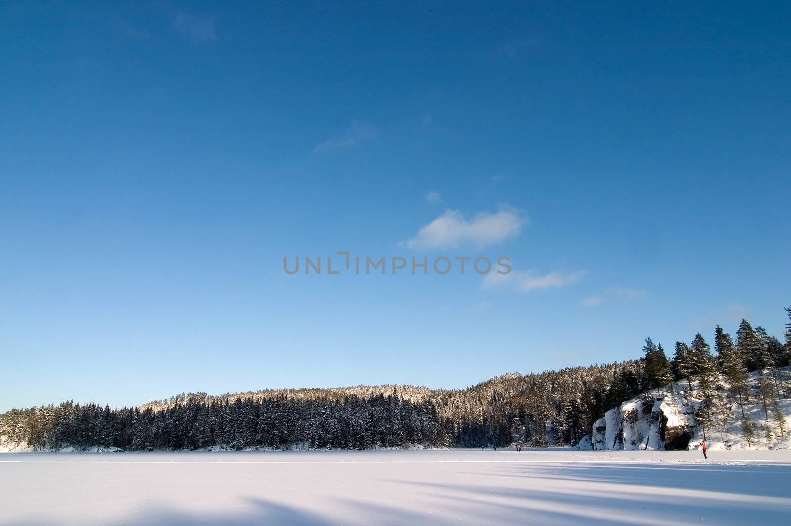 A frozen lake in the middle of winter on a beautfil blue sky day with a few people barely viewalbe skiing in the background.