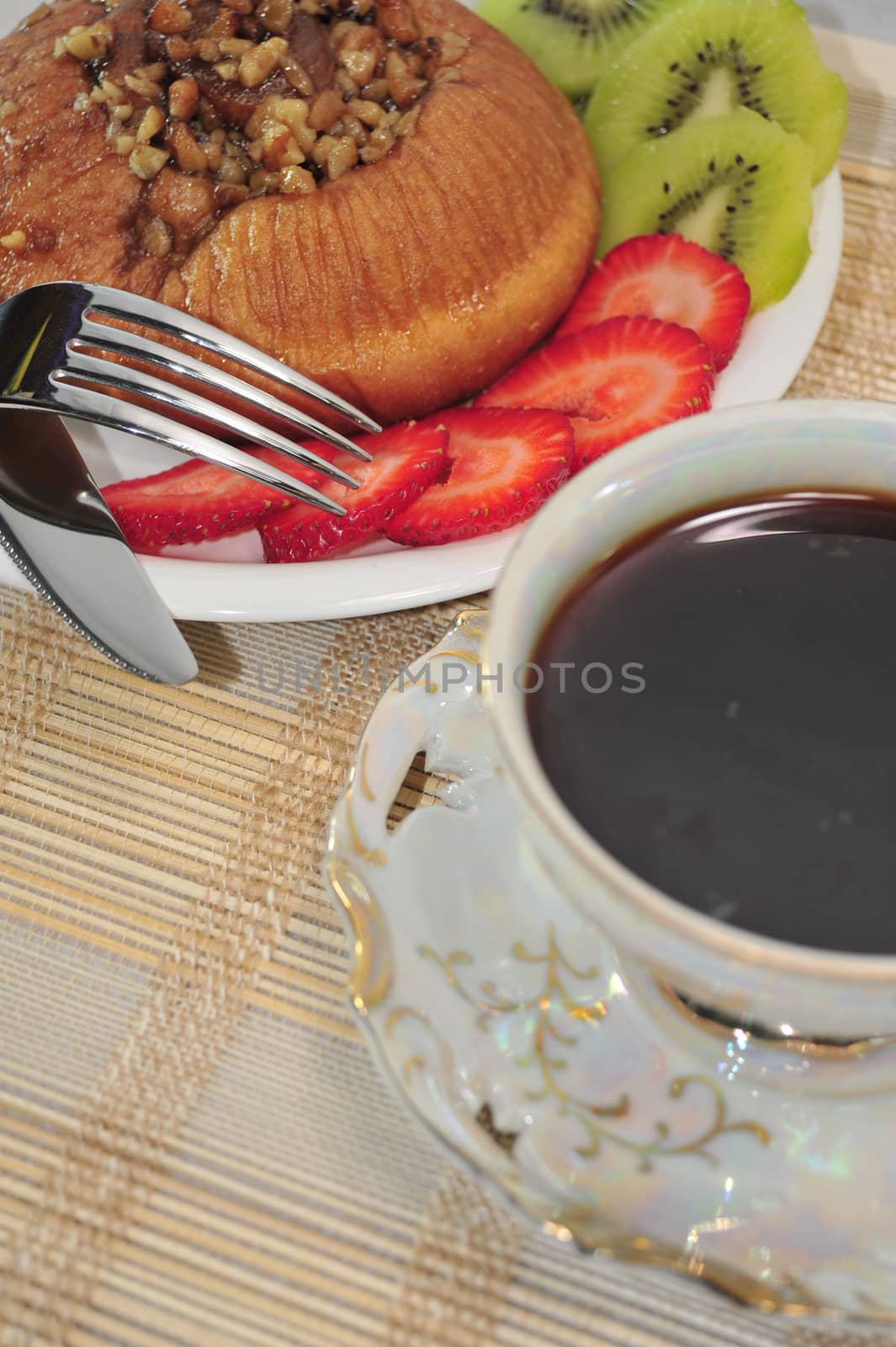 A fresh Cinnamon Roll topped with Walnuts, sliced Strawberries and Kiwi on the side and a hot cup of coffee.