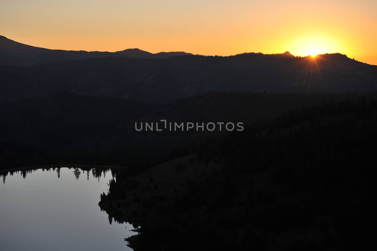 Darkness still keeps the mountains in shadow until the sun rises while the lake mirrors the surrounding trees.