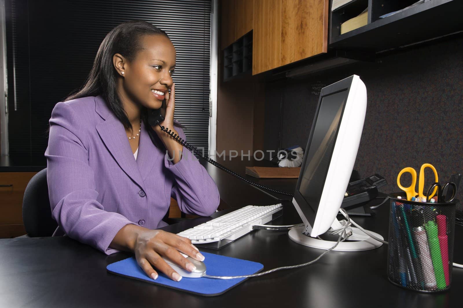 African-American young adult smiling business woman talking on phone and working at computer in office.